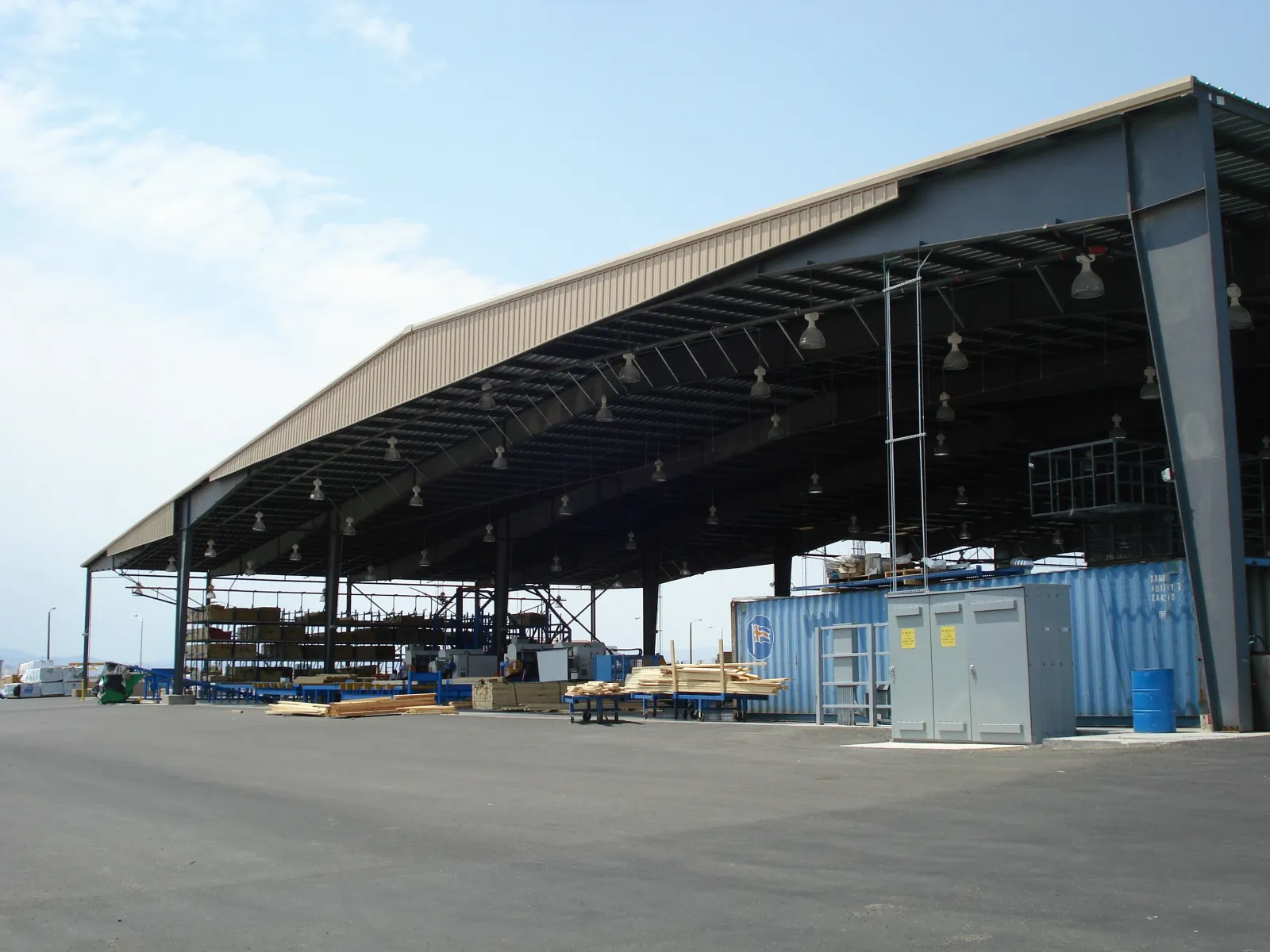 Large open warehouse structure with metal roof, containers, and industrial materials on concrete ground under clear sky