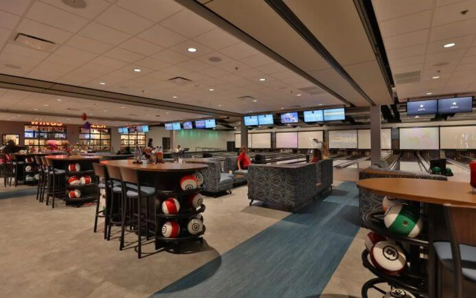 Modern bowling alley interior with seating, tables, bowling balls, and multiple lanes with screens.