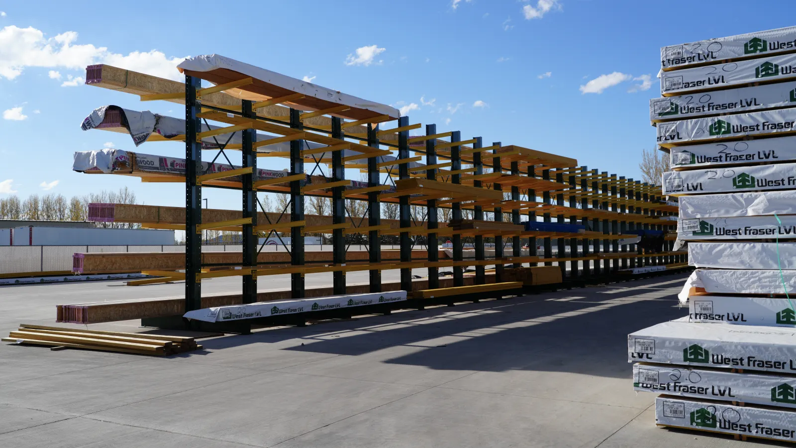 Outdoor lumber storage rack with long wooden beams and stacked West Fraser LVL packages on a sunny day.