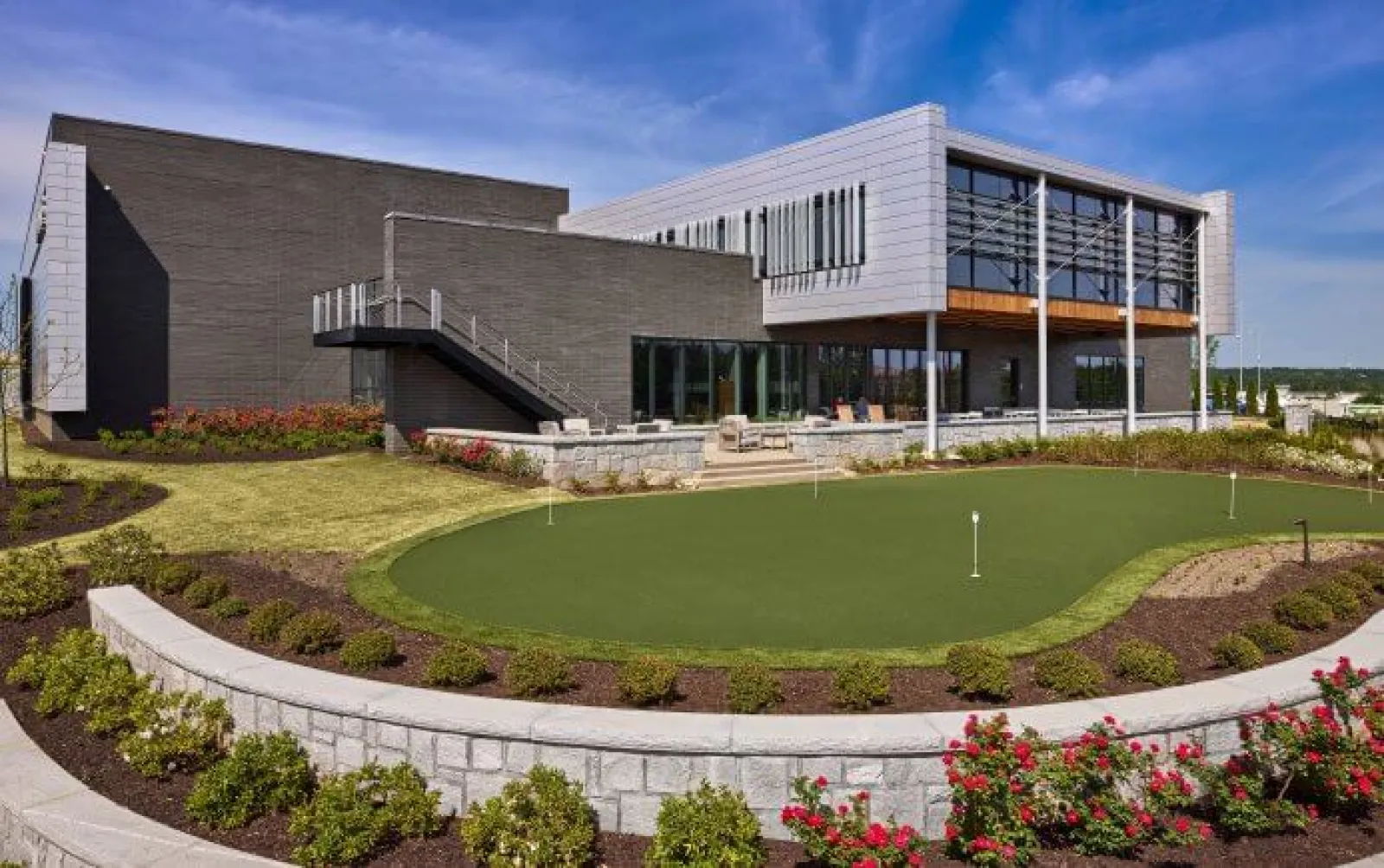 Modern building with large windows adjacent to a landscaped putting green surrounded by flower beds under blue sky