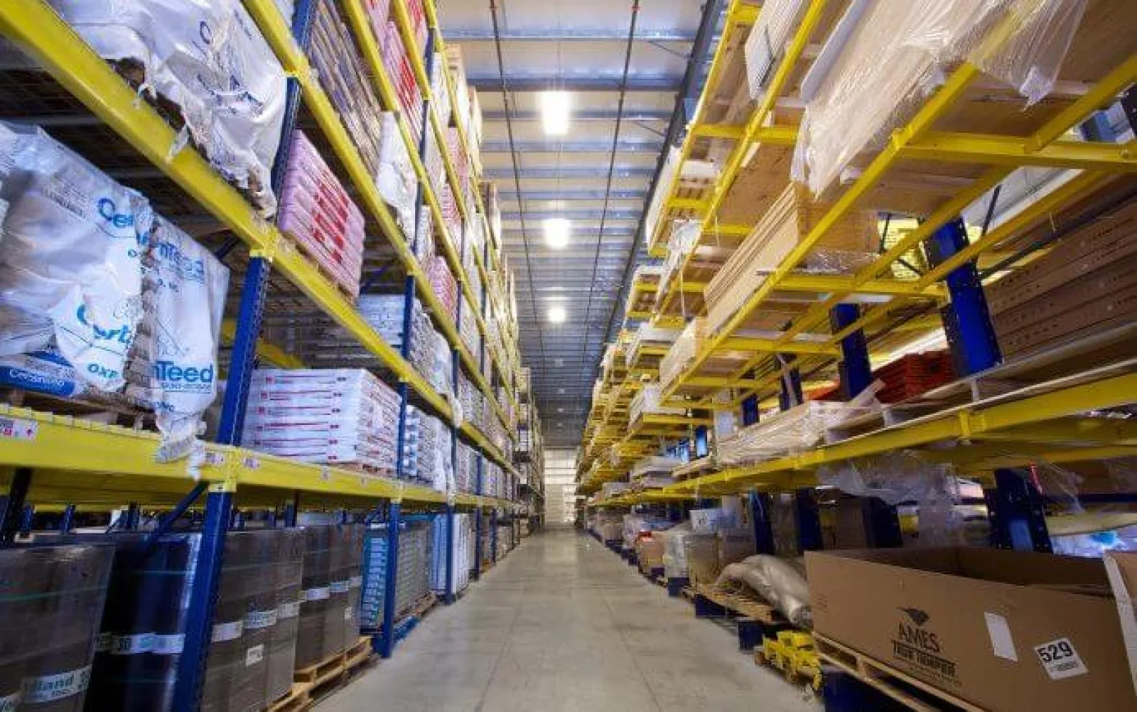 Wide aisle in warehouse lined with tall yellow shelves stocked with boxes and packaged goods.