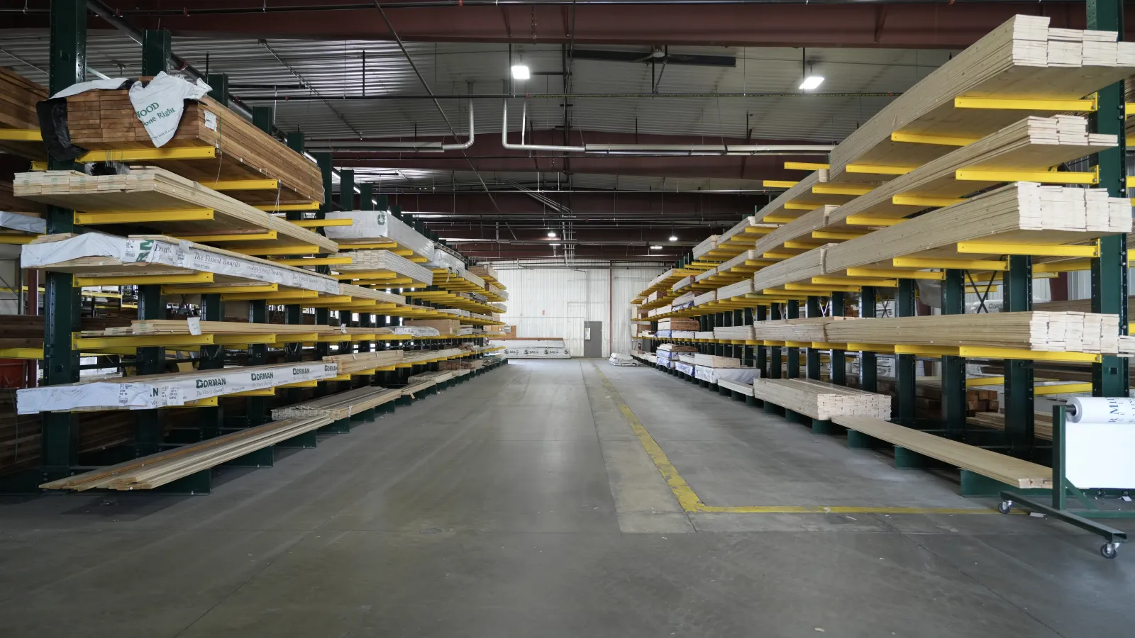 Interior view of a warehouse aisle with stacked lumber on metal racks and a concrete floor under overhead lighting.