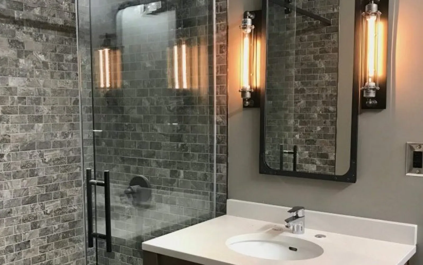Modern bathroom with glass shower, gray stone tiles, white countertop, and industrial wall light fixtures.