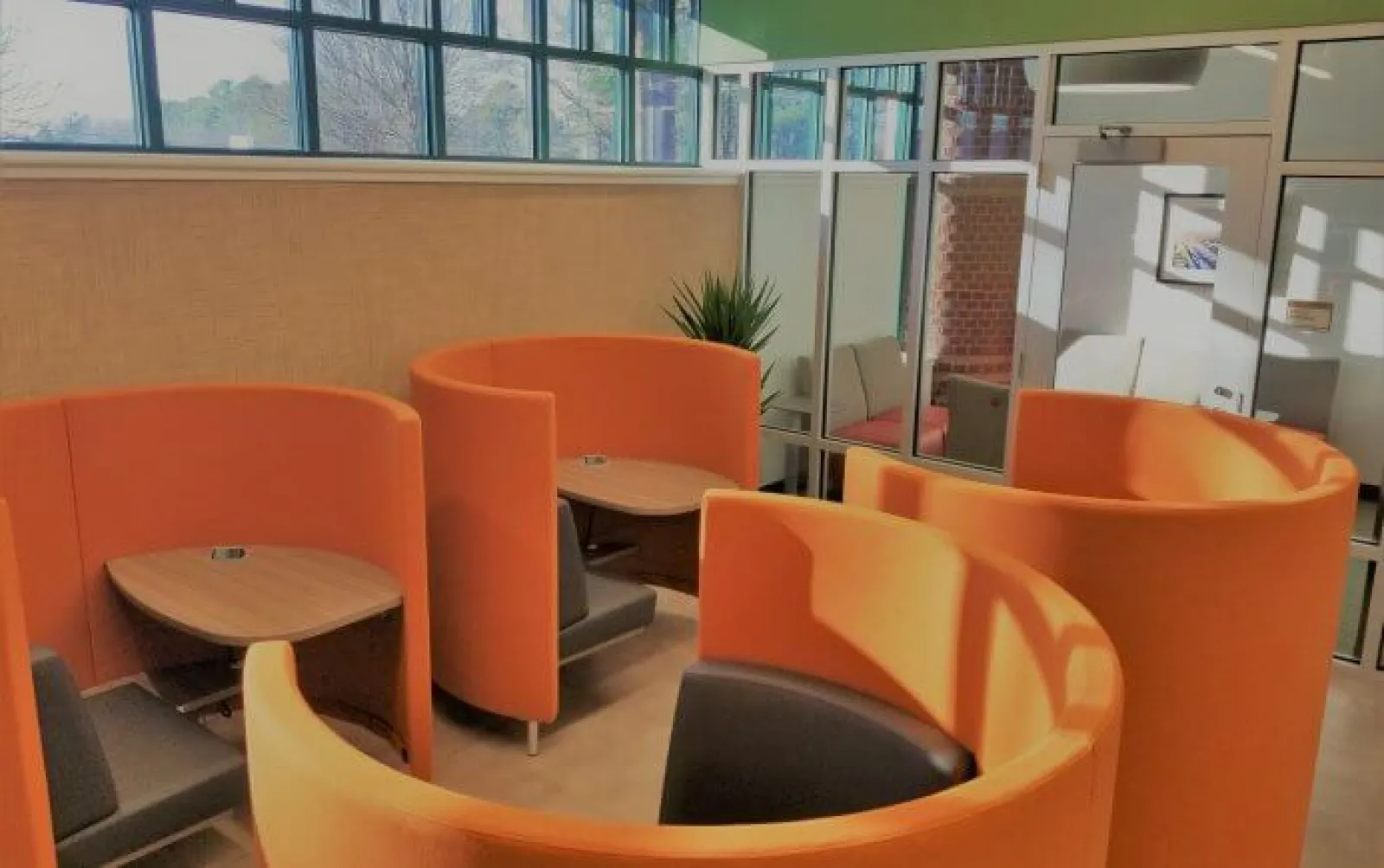 Modern office lounge area with curved orange seating pods, small wooden tables, and black chairs under natural light.