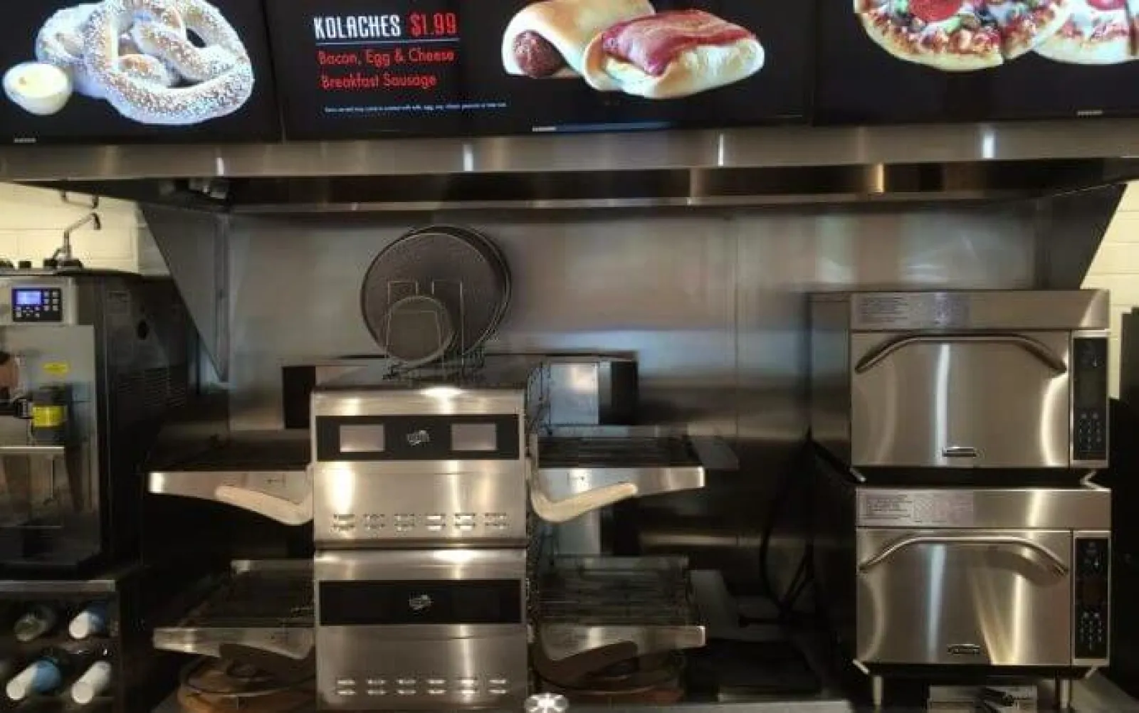 Commercial kitchen with stainless steel pizza ovens, sandwich press, and menu boards displaying breakfast items.