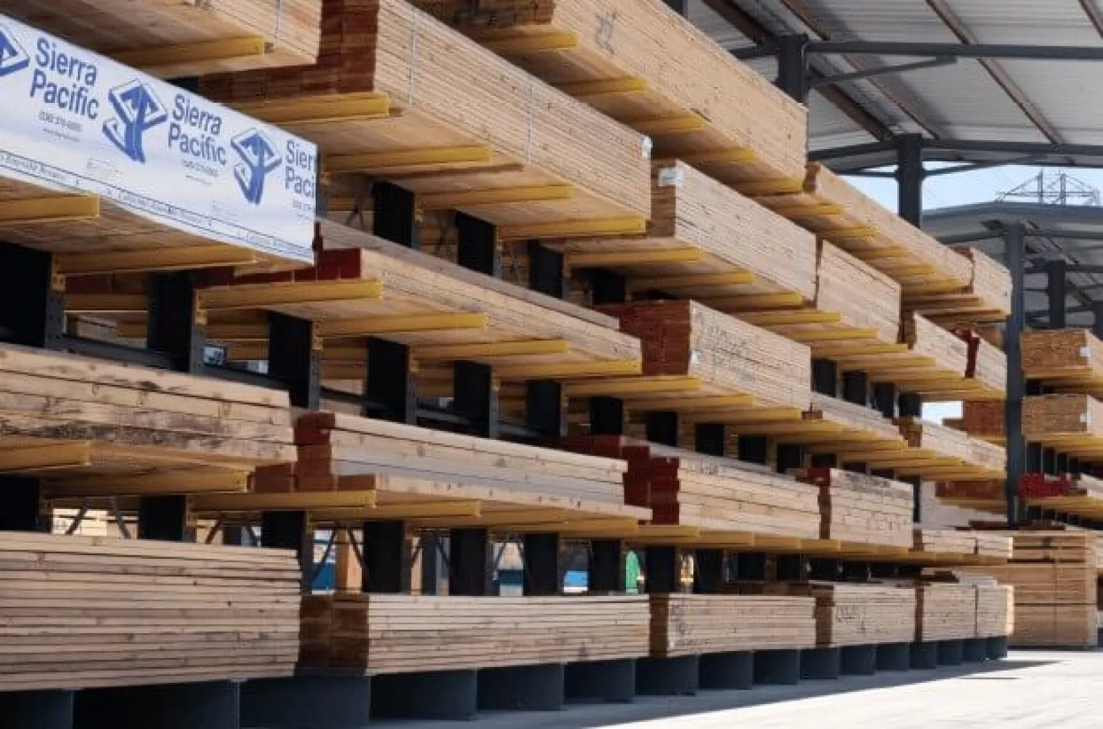 Stacks of lumber stored on metal racks at an outdoor lumber yard under a metal roof structure.