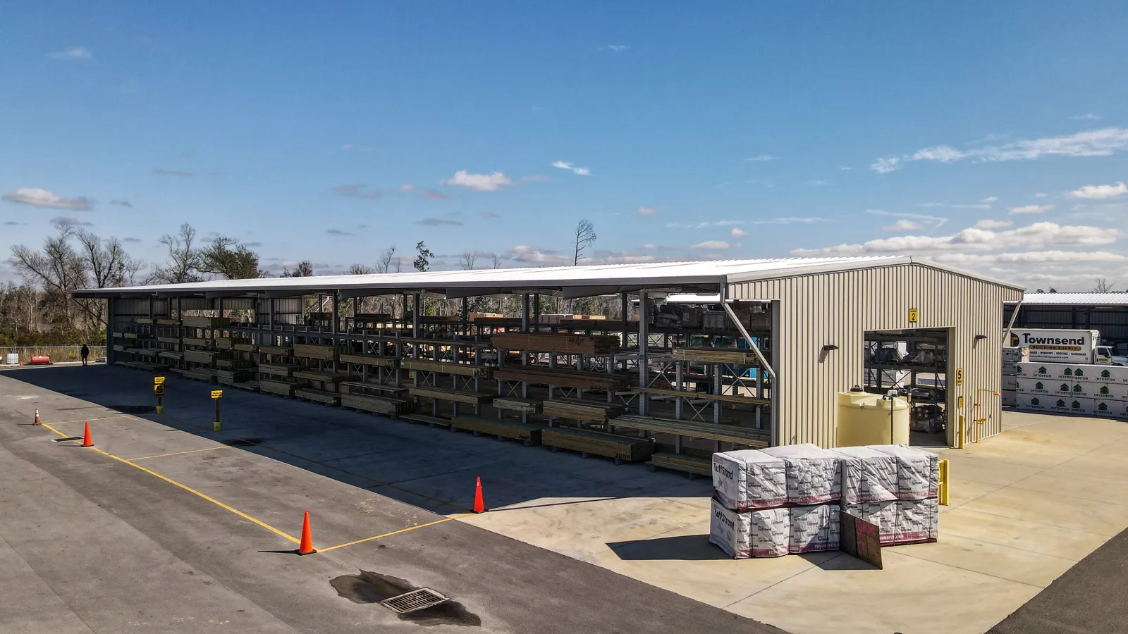 Outdoor lumber storage rack under metal shelter with pallets and an industrial warehouse in a sunny setting.