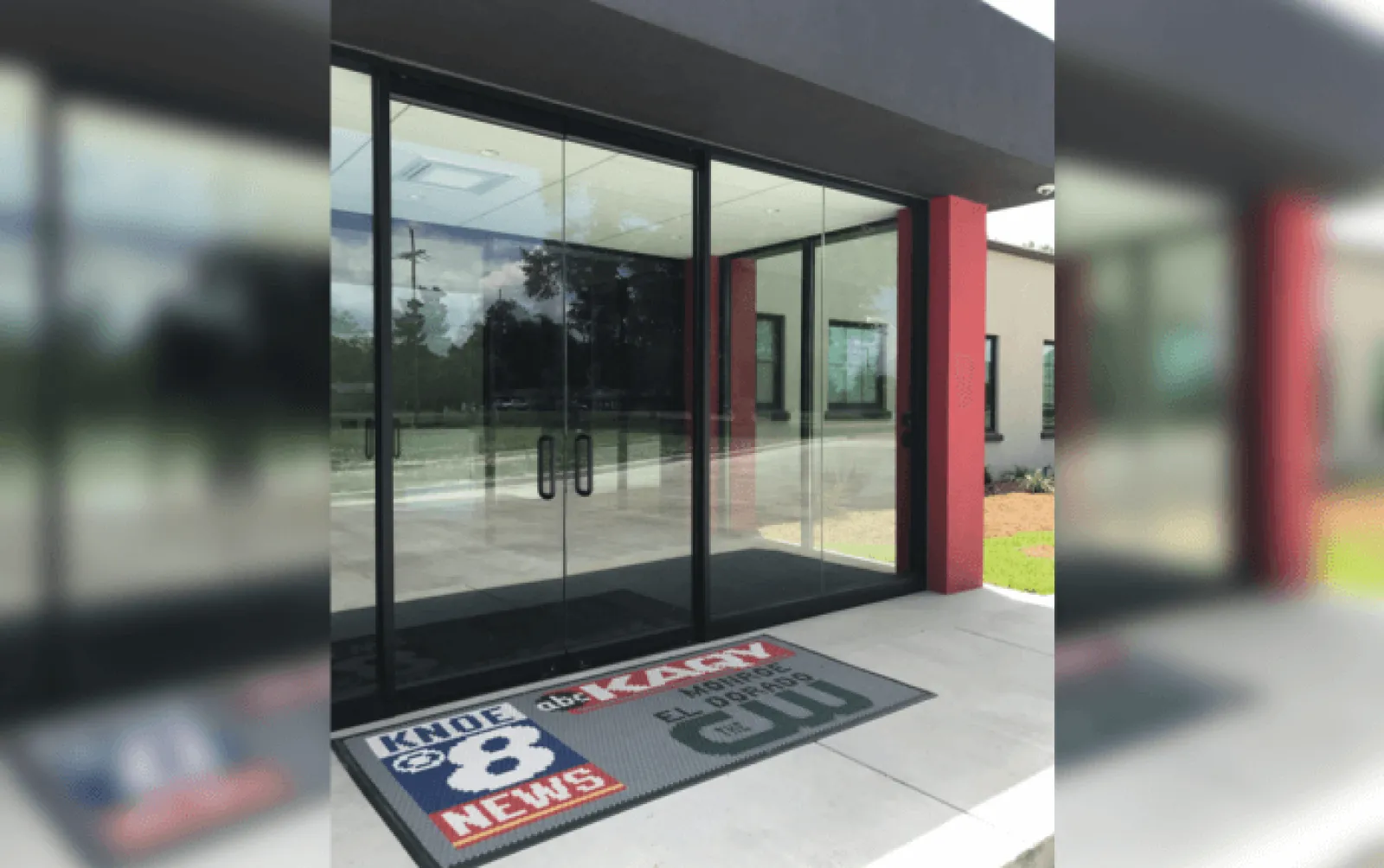 Glass entrance doors with KNOE 8 News and local TV station logos on the floor mat outside a building.