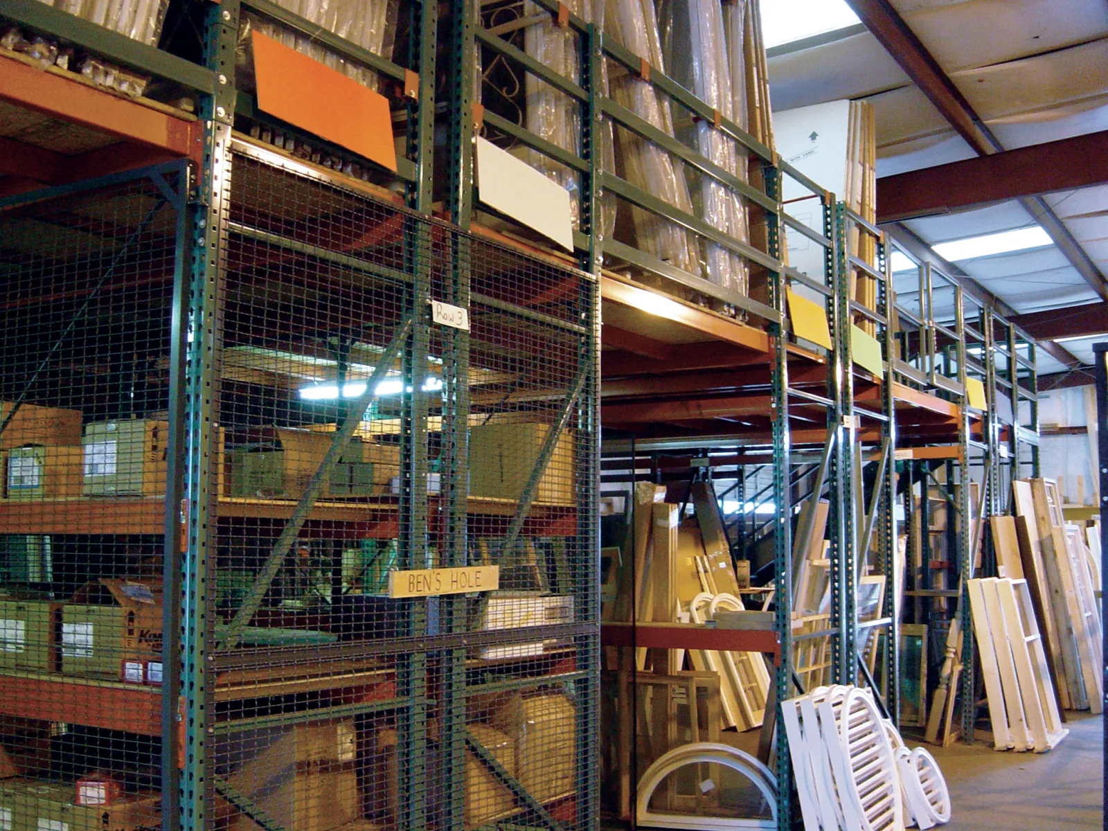 Warehouse storage with metal shelving holding boxes and wooden panels under industrial roof lighting.