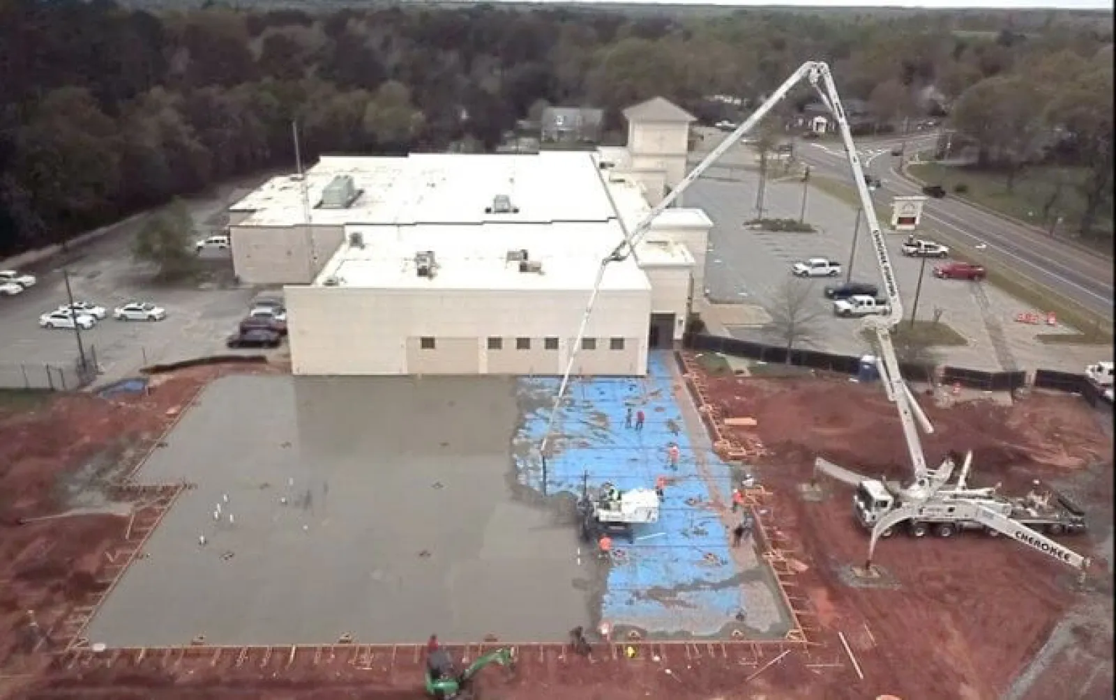 Aerial view of concrete pouring at construction site next to a white building and parking lot with trees in the background