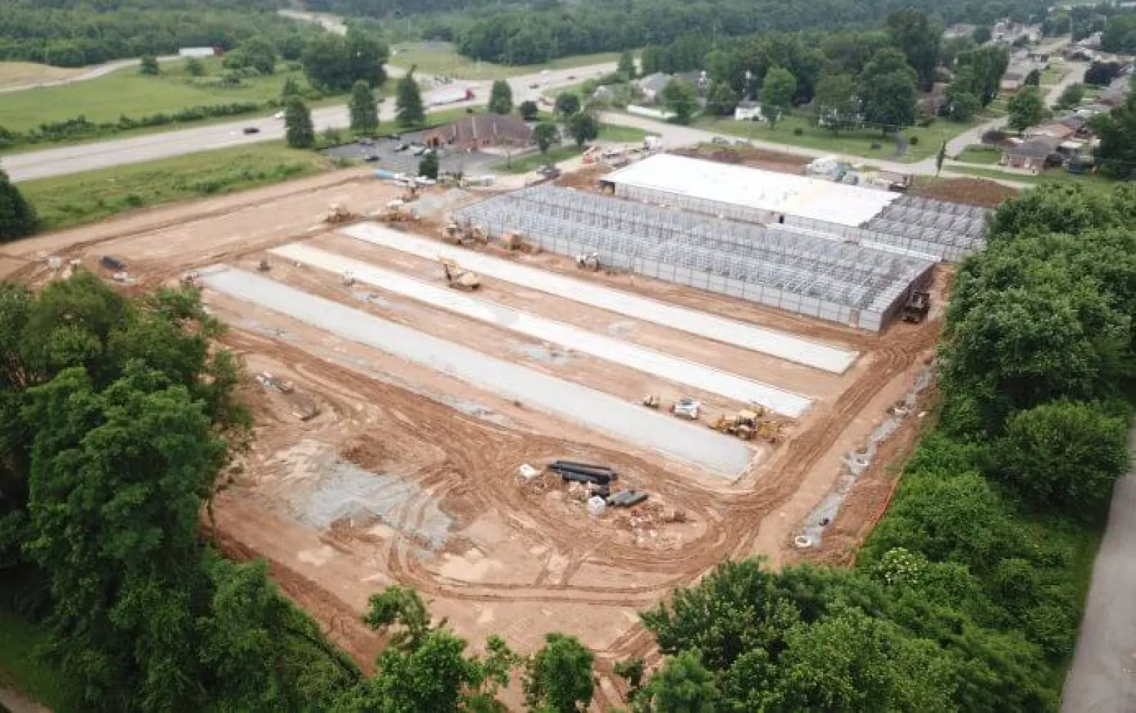 Aerial view of a construction site with foundation slabs and steel framework surrounded by greenery and nearby roads.