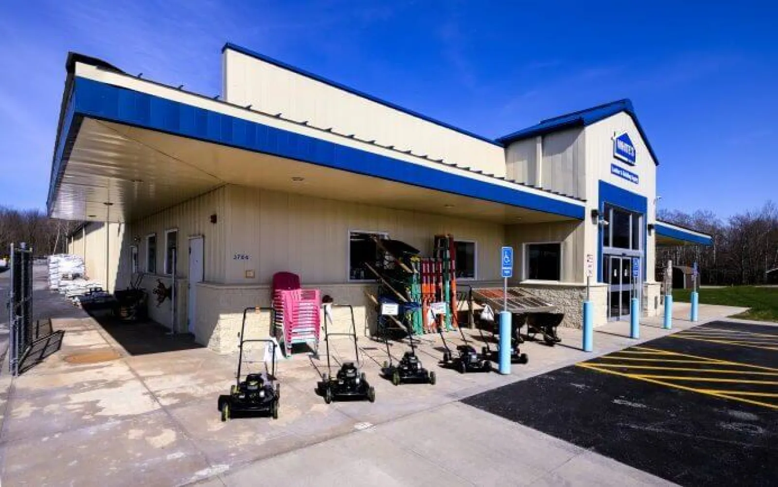 Outdoor picture of a hardware store with lawnmowers and garden tools displayed on the sidewalk under a blue awning.