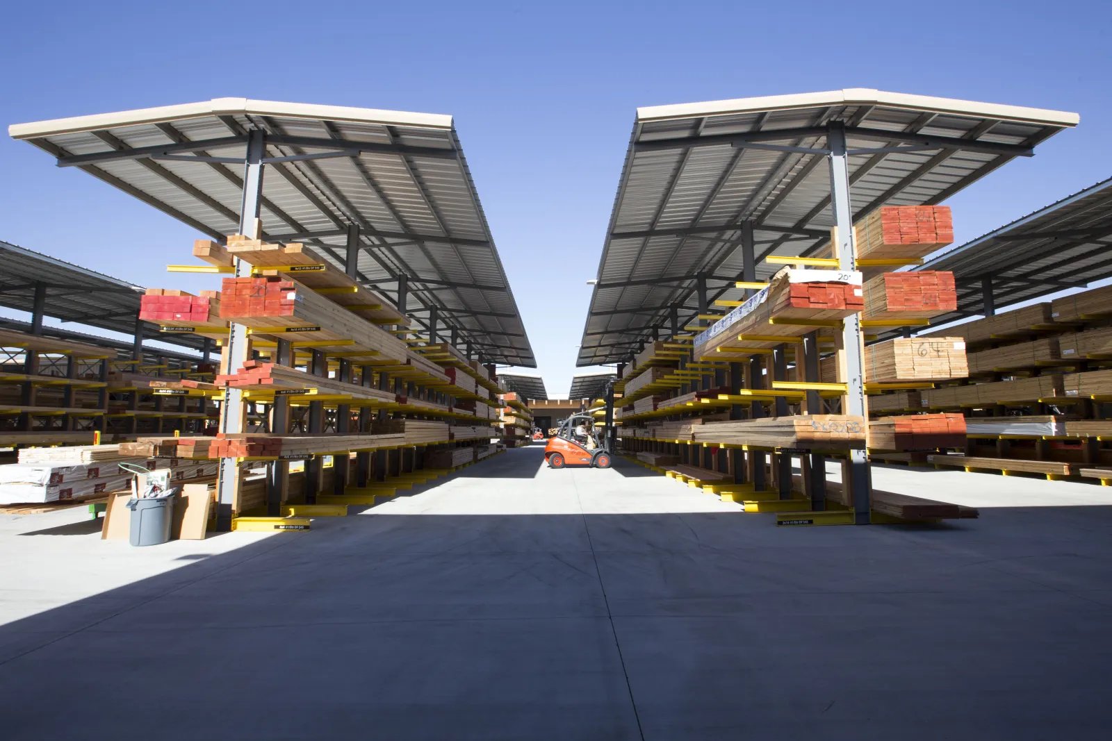 Outdoor lumber yard with organized stacks of wooden planks under metal roofs and a forklift in the distance.