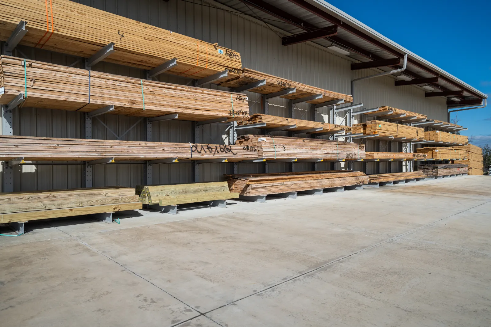 Outdoor lumber storage racks holding various types of wood planks under clear blue sky at a hardware store.