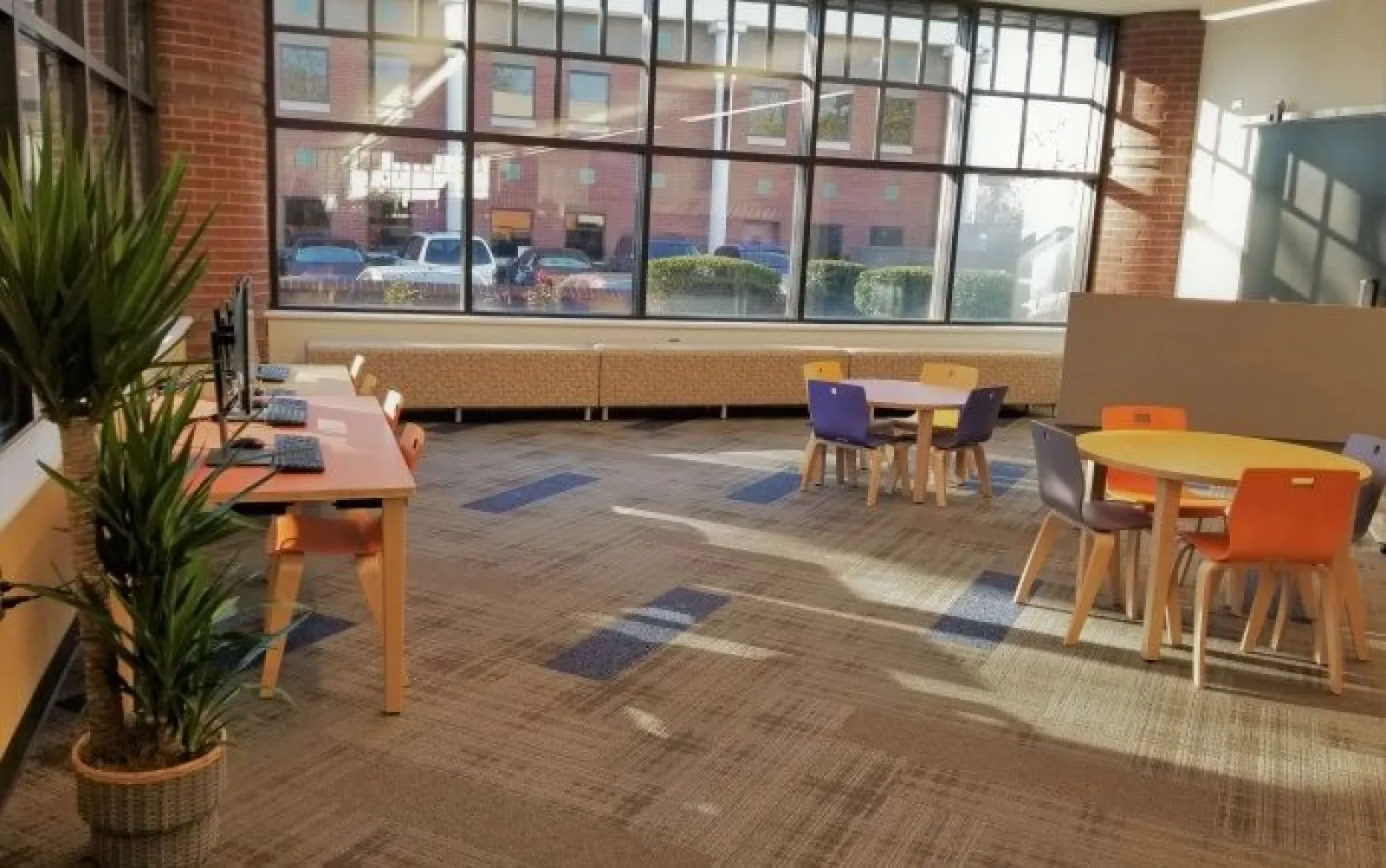 Bright modern workspace with computers, colorful chairs, round tables, large windows, and a potted plant.