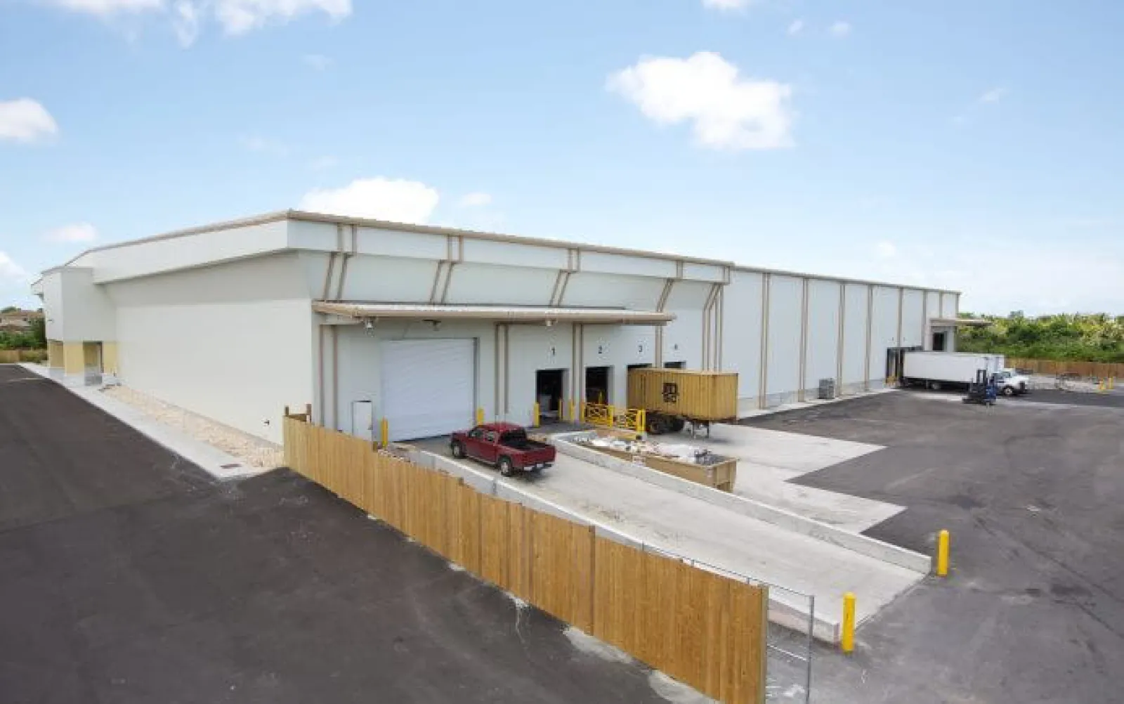 Modern warehouse with loading docks, trucks, and fenced parking area under a blue sky.