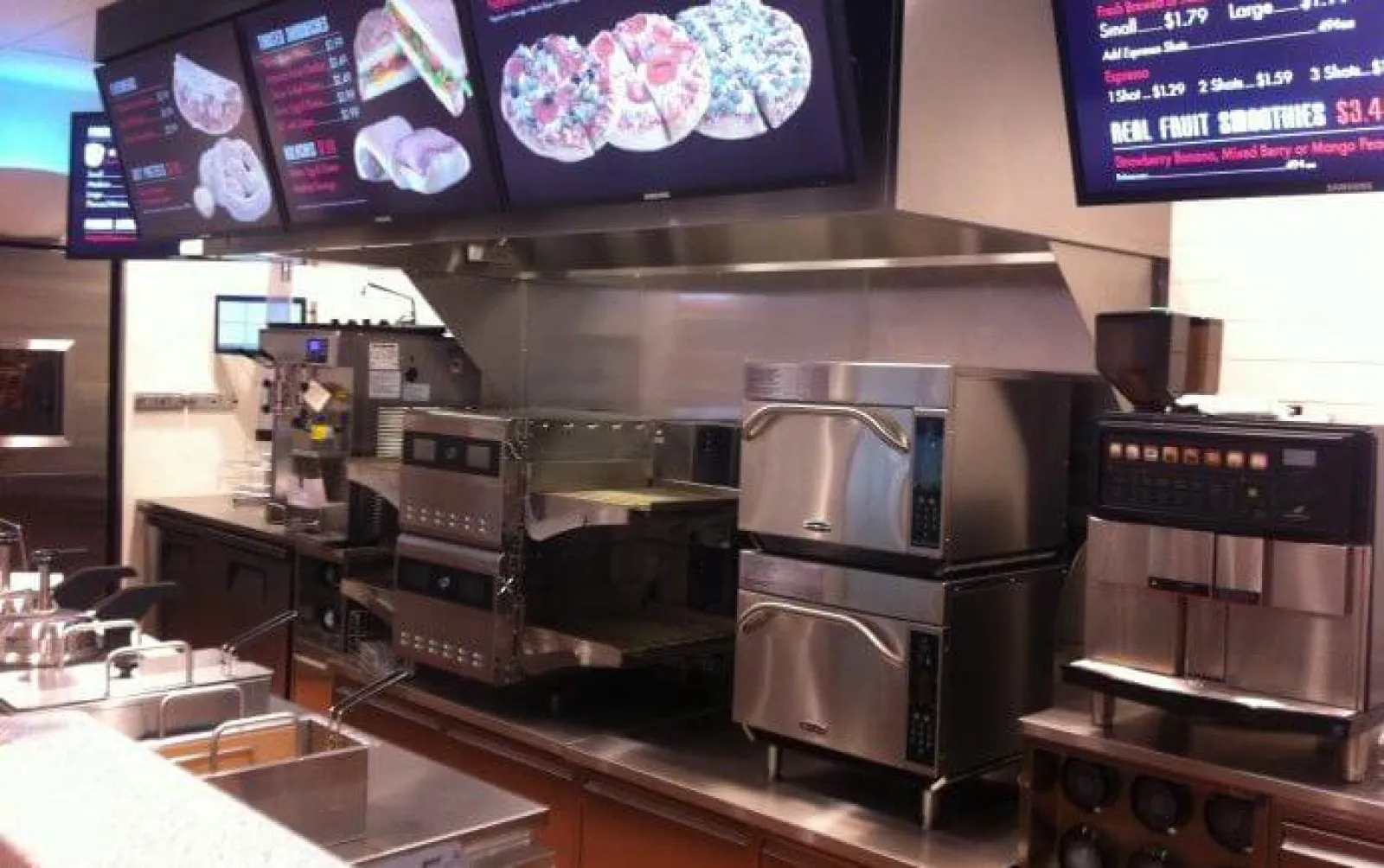 Modern pizza restaurant kitchen with digital menu boards and stainless steel ovens and equipment.
