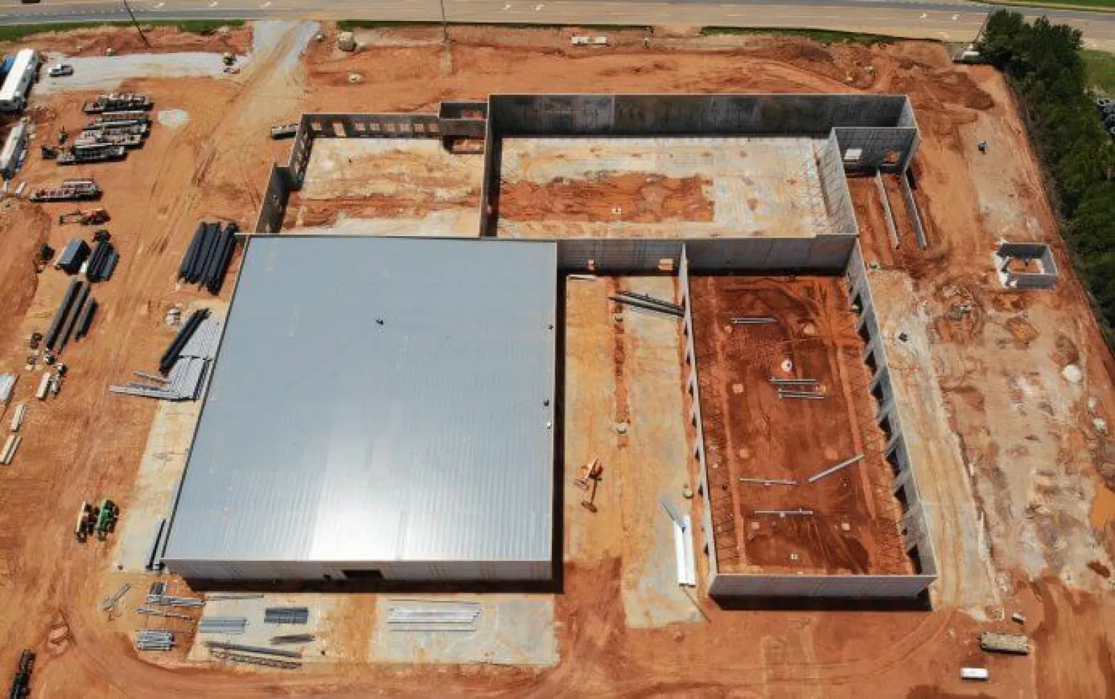 Aerial view of a construction site with a large metal-roofed building and adjacent concrete foundation walls on red soil.