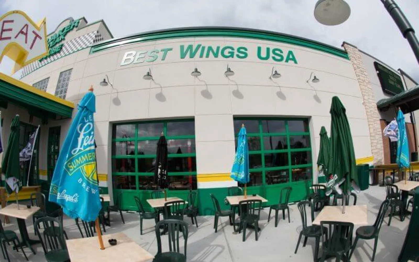 Outdoor seating area at Best Wings USA restaurant with green chairs, tables, and colorful umbrellas on a sunny day.