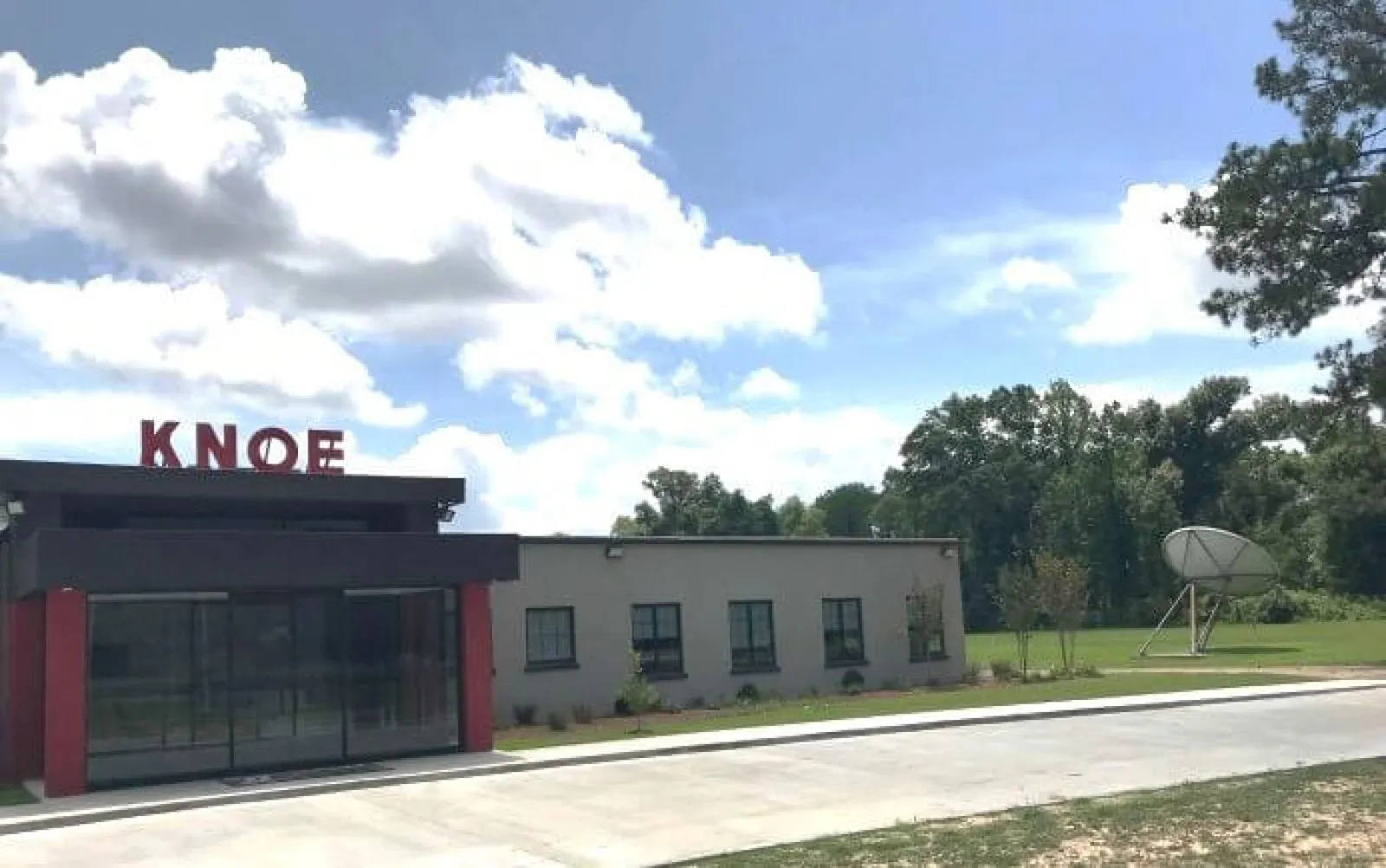 Exterior view of a small building with KNOE sign on the roof under a blue sky with clouds and surrounding greenery.