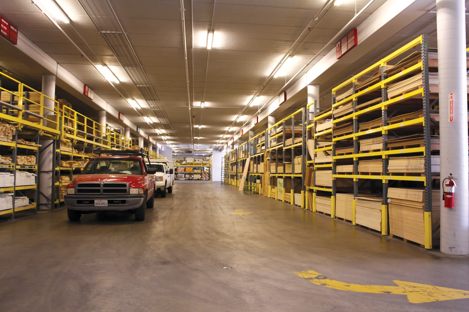 Interior of a spacious warehouse with wooden boards on yellow racks and two parked pickup trucks