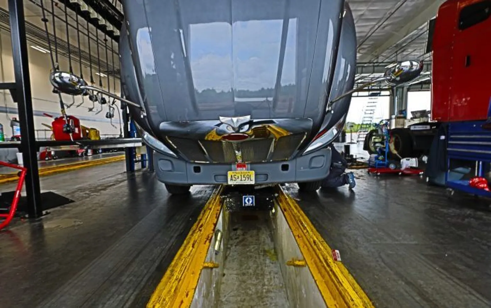 Front view of a truck with lifted hood in a maintenance garage over a service pit with mechanic working underneath.