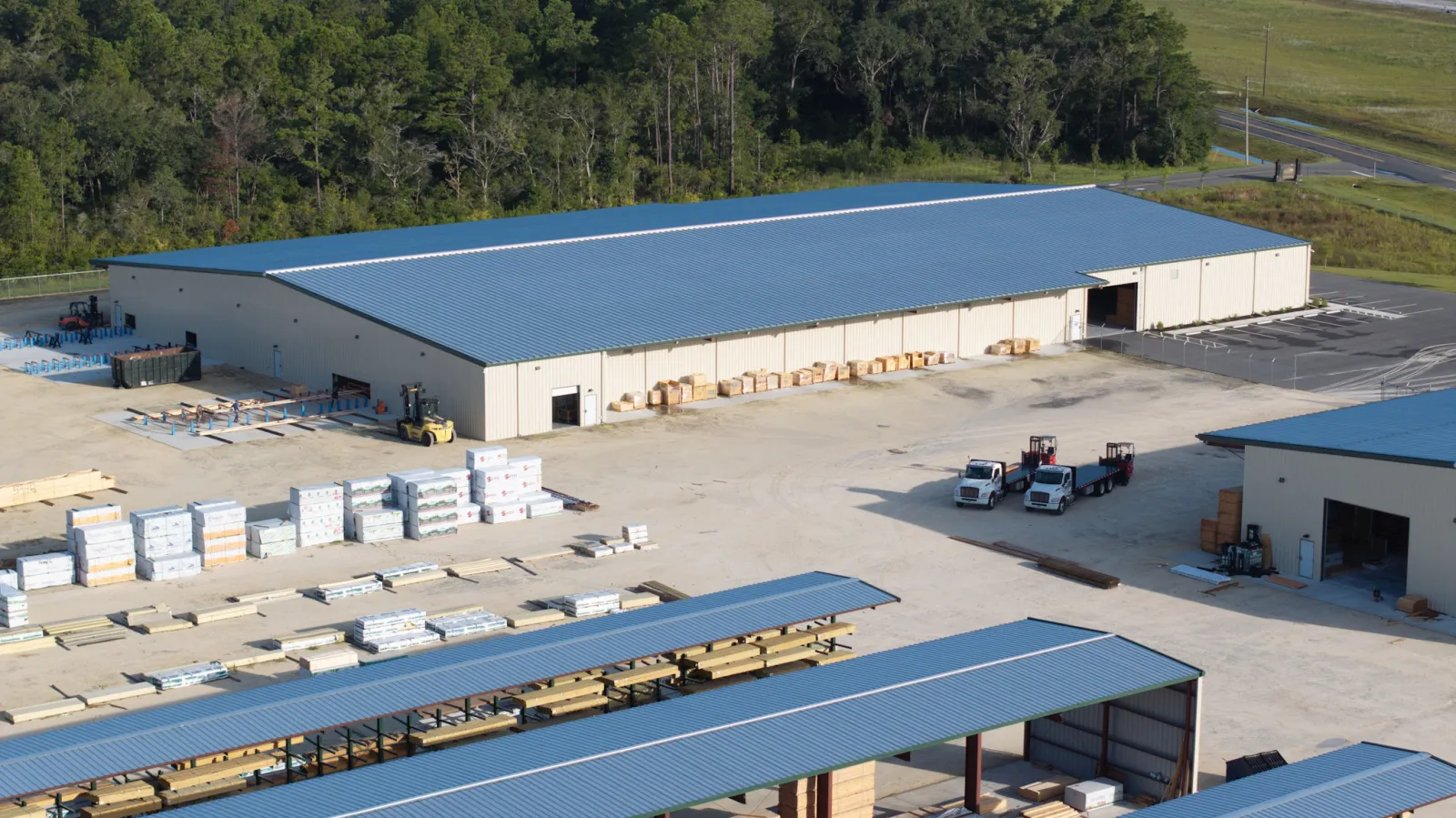 Aerial view of a lumberyard with stacked wood, large storage buildings, and trucks under blue roofs.