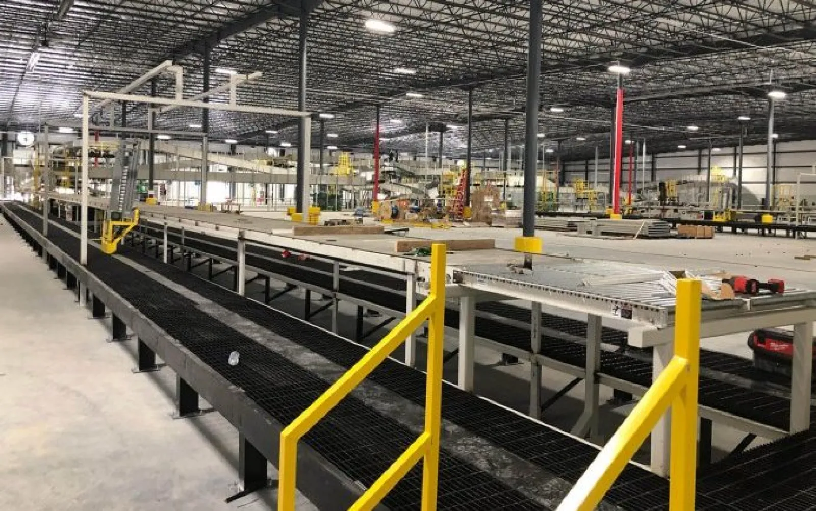 Interior of a large industrial warehouse with conveyor belts and yellow safety railings under metal roof lights.
