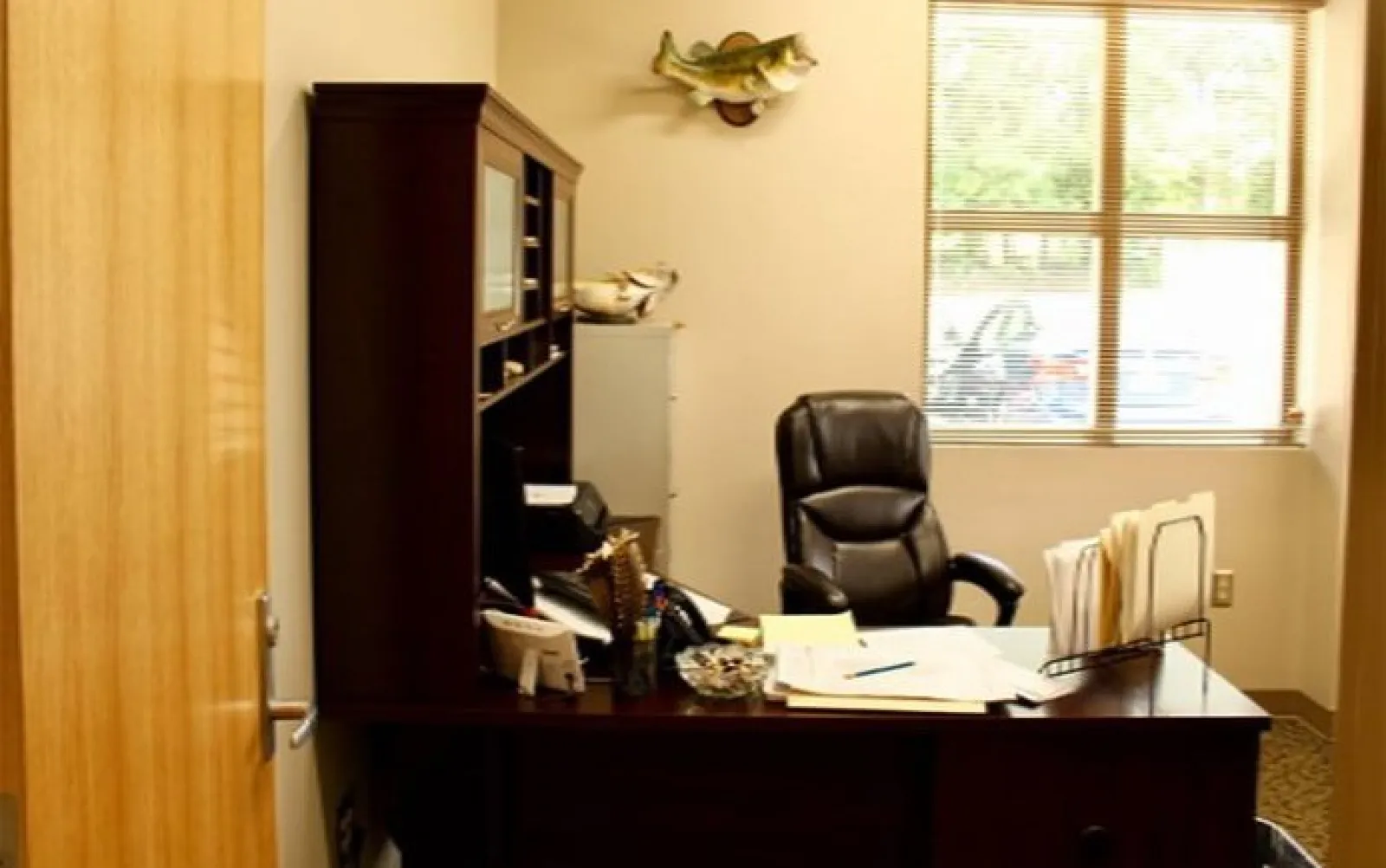 Cozy office setup with dark wood desk, leather chair, paperwork, filing system, and a fish mounted on the wall.