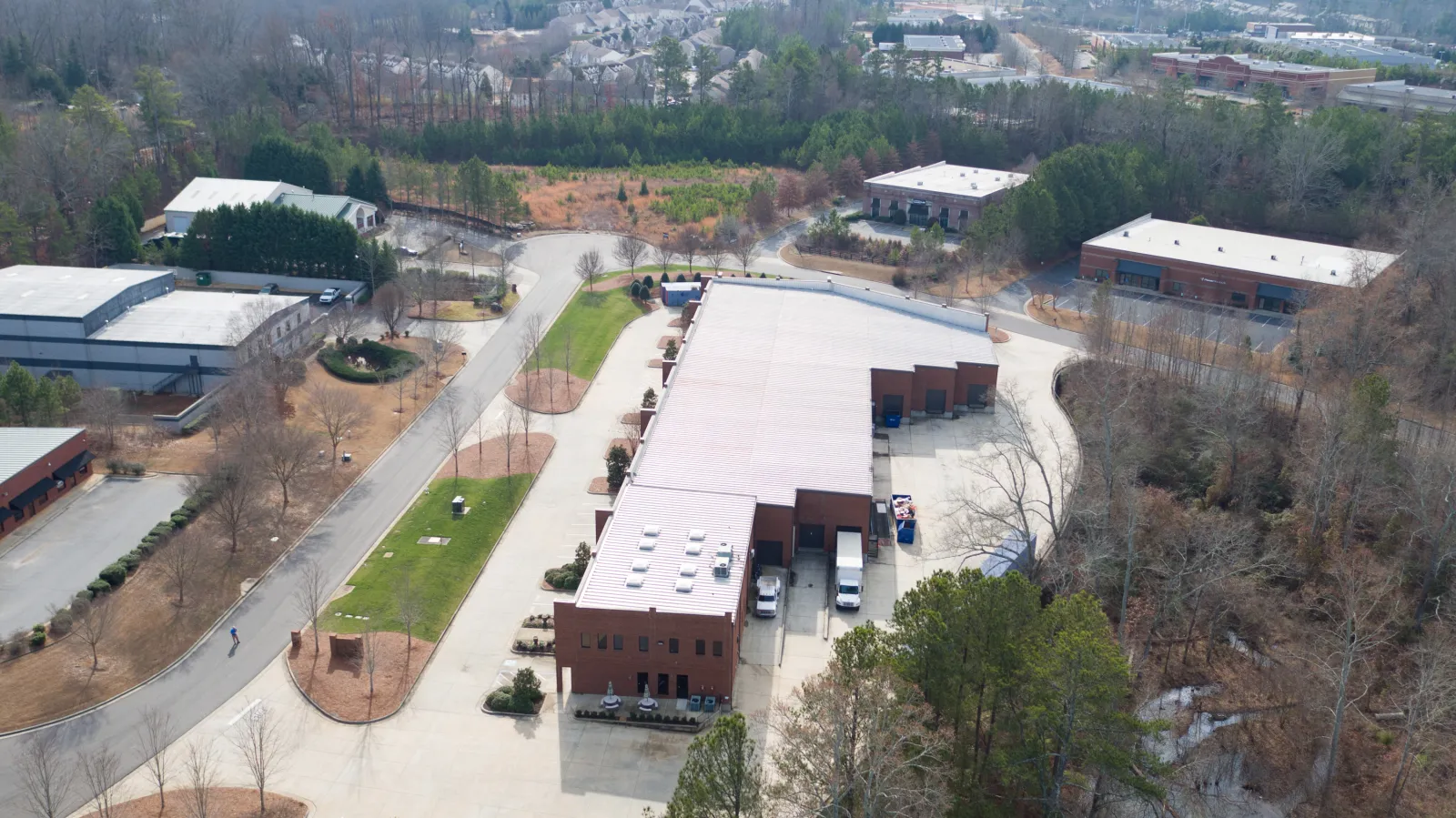 Aerial view of a large industrial warehouse complex surrounded by trees and nearby residential area under clear sky.