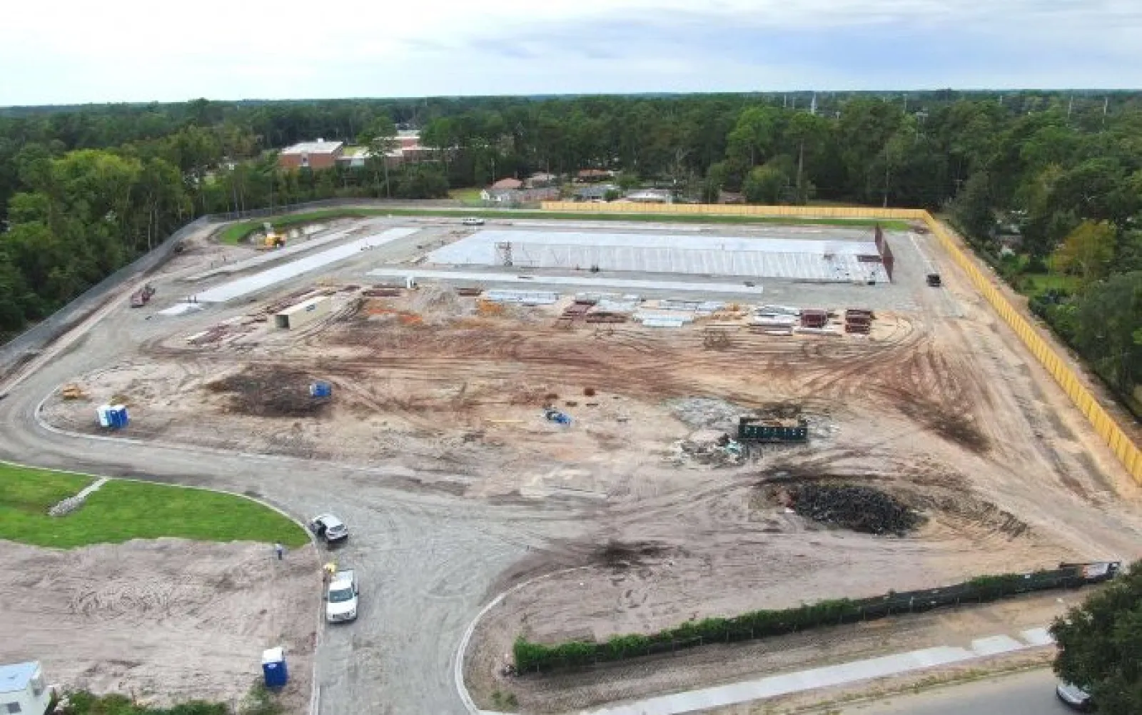 Aerial view of a large construction site with earthworks, concrete slabs, equipment, and surrounding trees under a cloudy sky