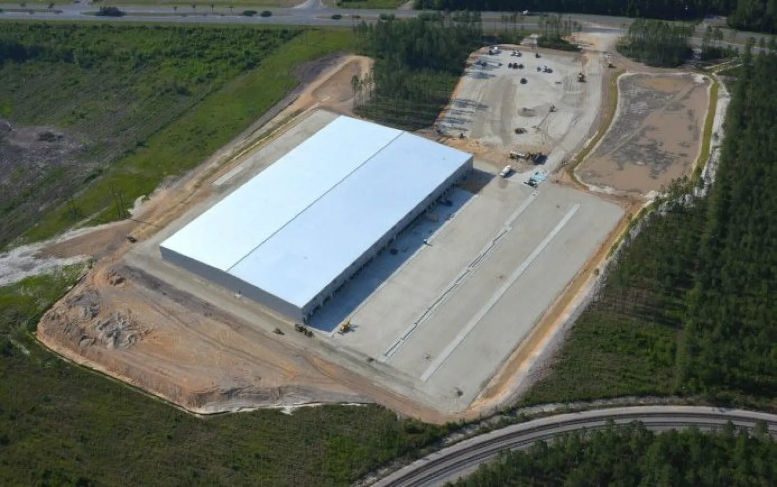 Aerial view of a large white industrial warehouse surrounded by cleared land and greenery with adjacent roads.