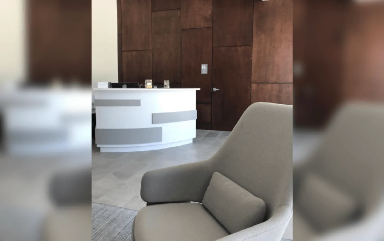 Modern reception area with a curved white desk, gray armchair, and wooden paneled wall with door.