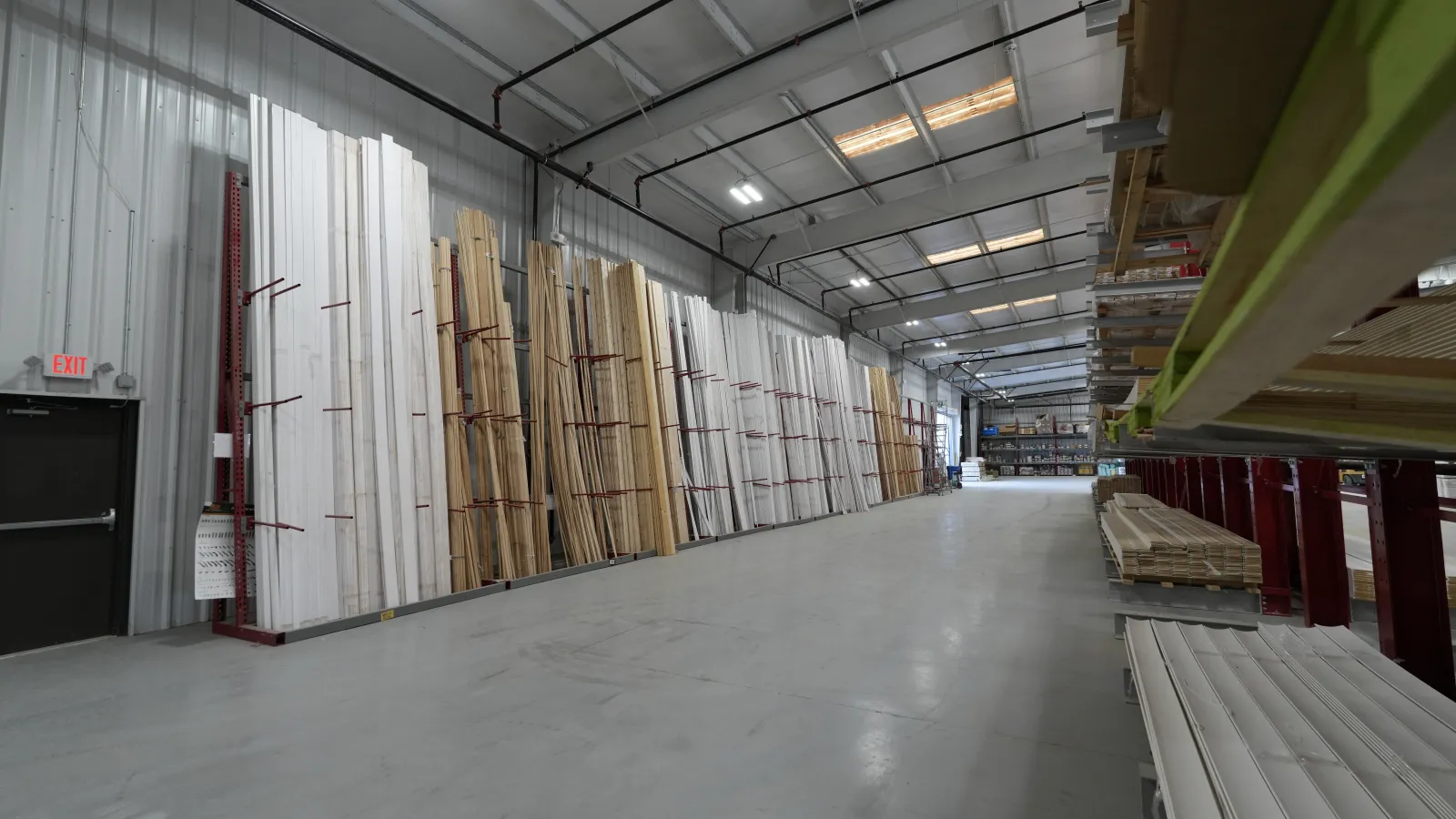 Interior view of a lumber warehouse with stacked wood panels and planks on racks under bright lighting.