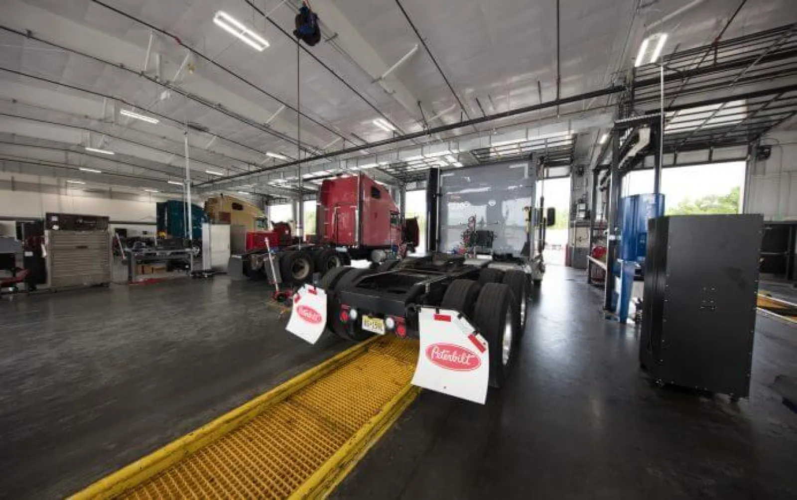 Inside a large truck repair shop with semi-trucks and maintenance equipment under bright lights.