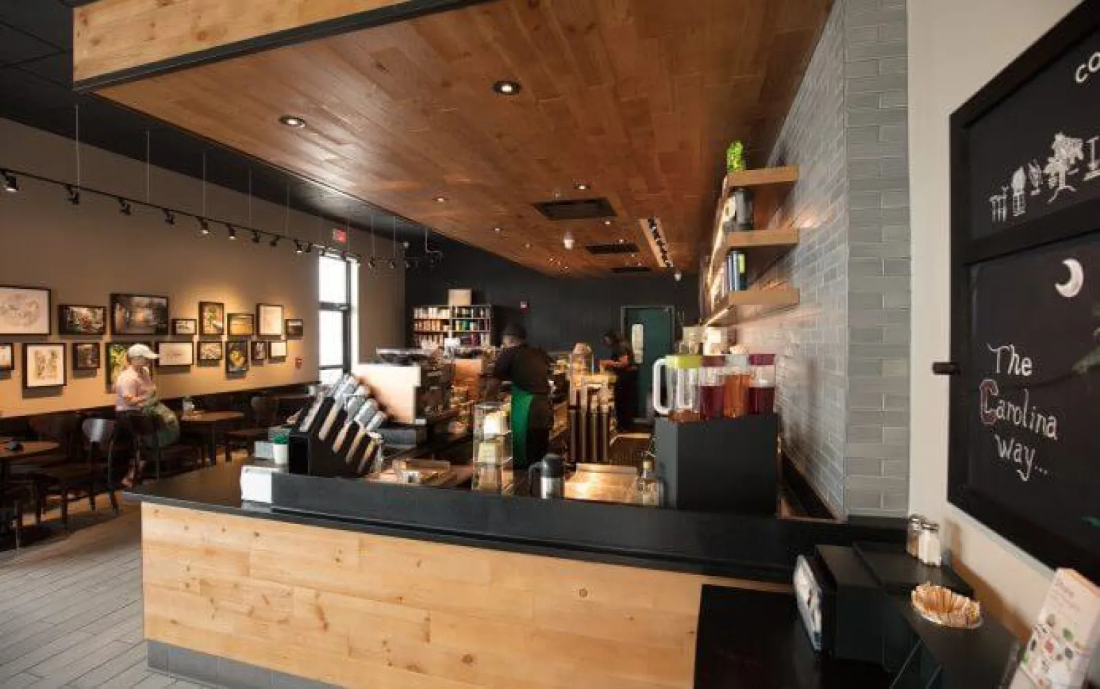 Cozy modern coffee shop interior with wooden accents, barista at counter, and customers seated by framed artwork.