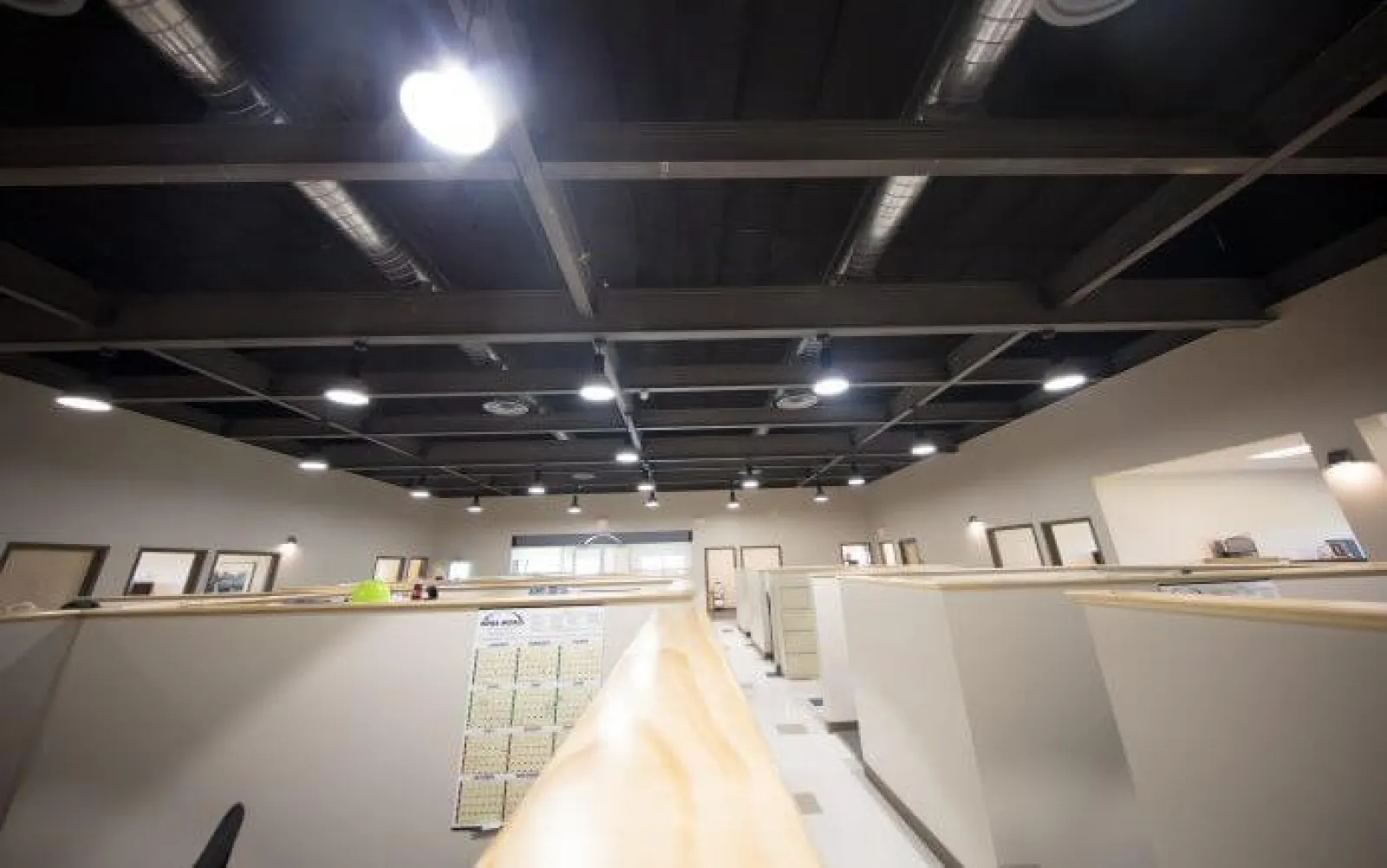 Modern office interior with beige cubicles, exposed black ceiling, and bright overhead lighting.