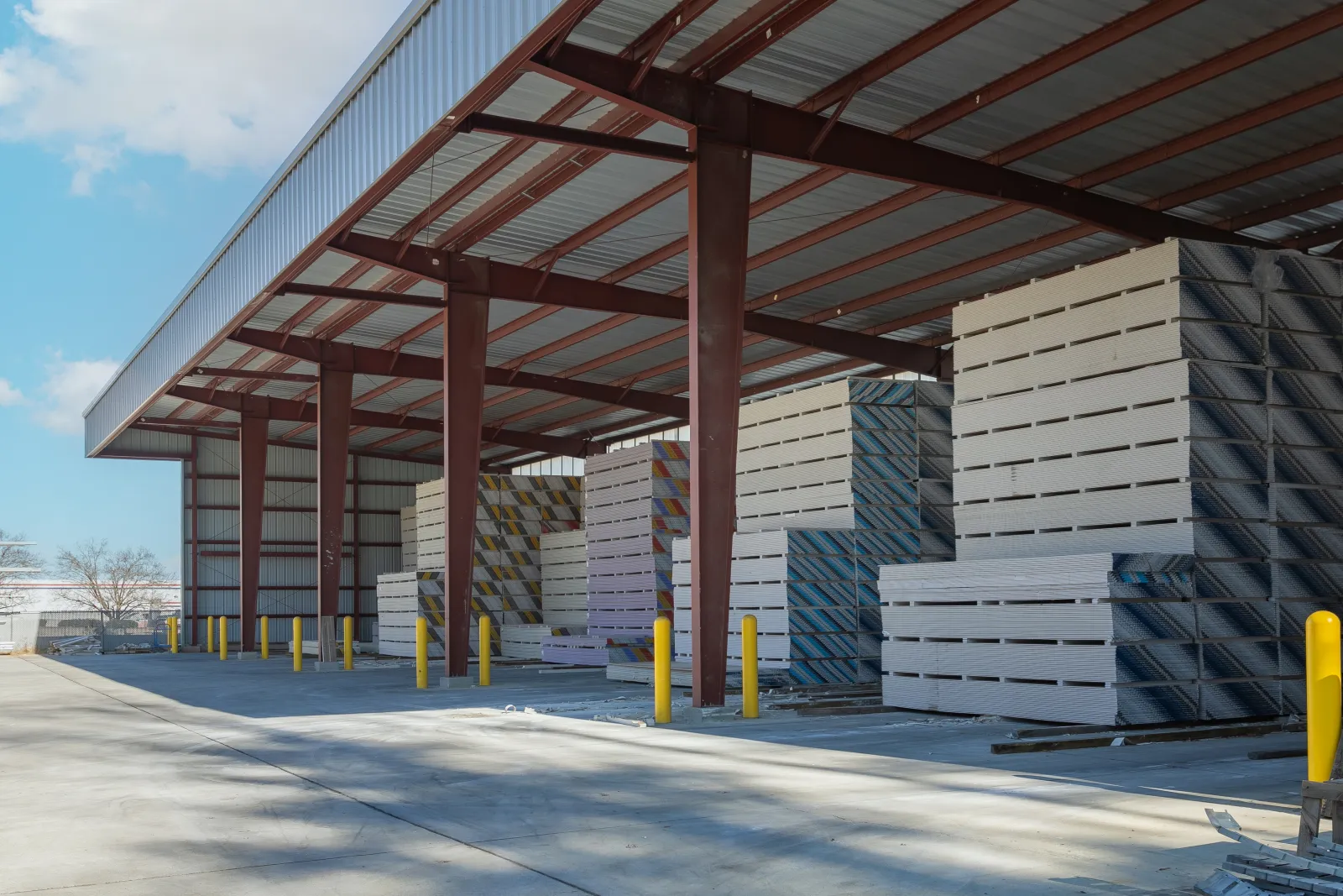 Stacks of lumber stored under a large open metal shelter on a sunny day with blue sky.