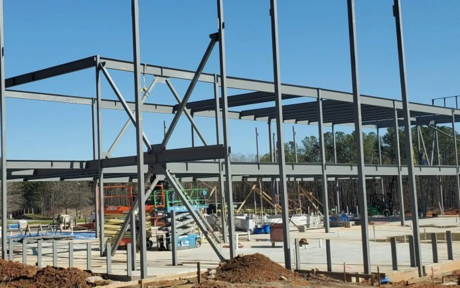 Steel framework of a building under construction on a clear sunny day with construction equipment and dirt piles.