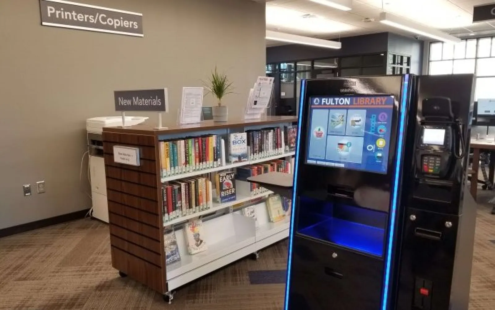 Library book return kiosk next to bookshelves of new materials and a printers/copiers station.