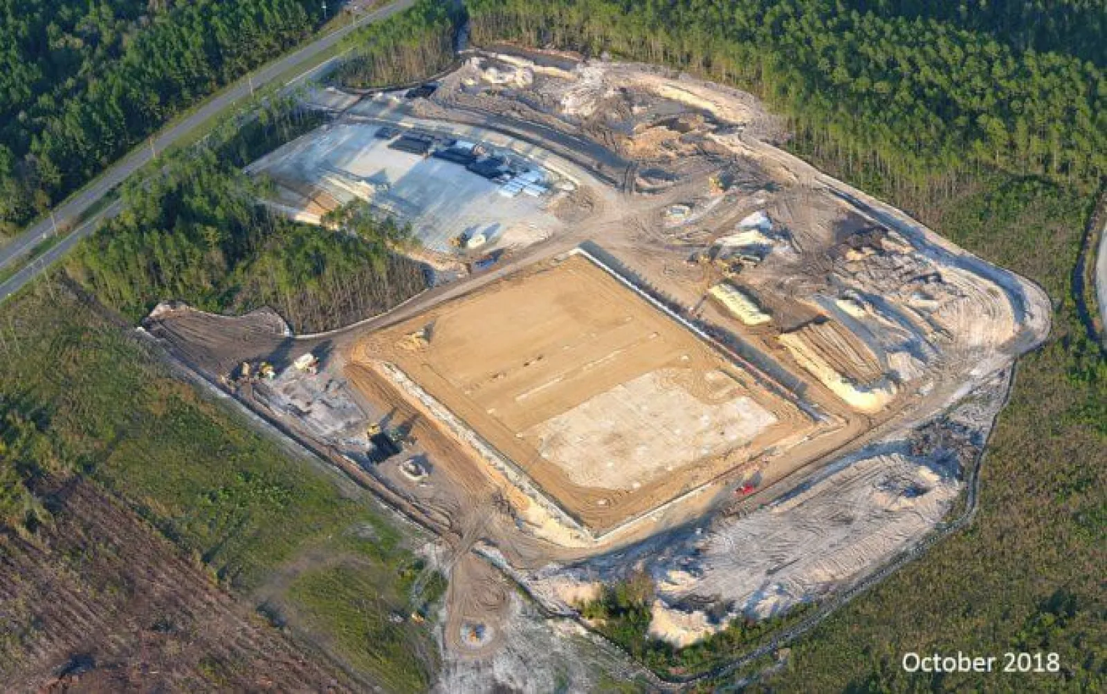 Aerial view of a large cleared construction site surrounded by forested land with groundwork in progress.