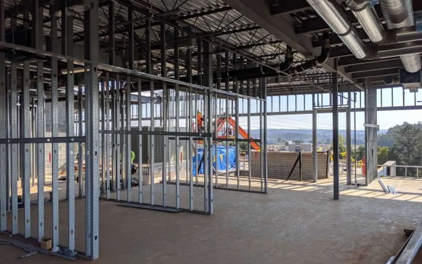 Interior construction site with metal framing, exposed ceiling ducts, and large windows overlooking outdoor scenery.