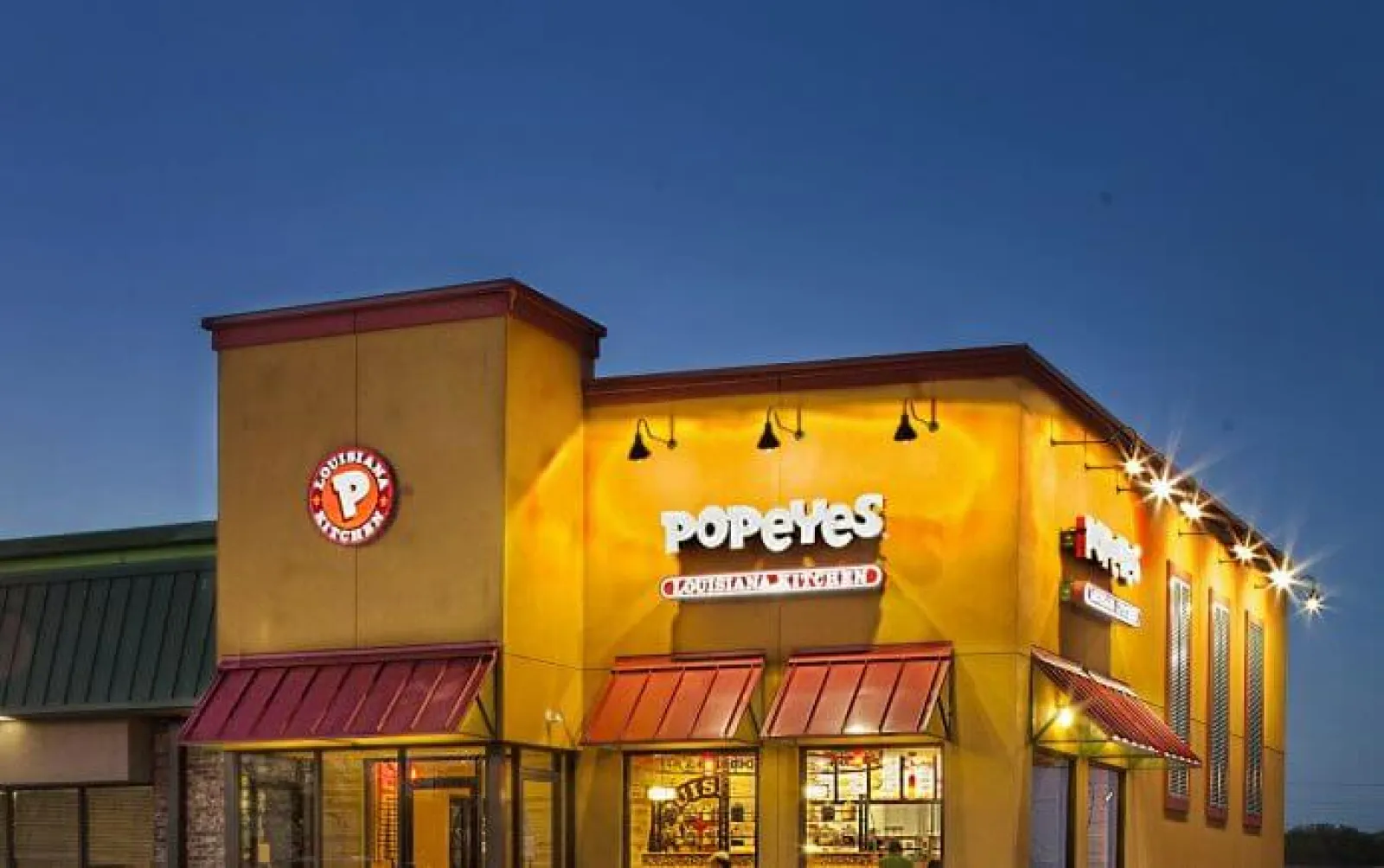 Popeyes Louisiana Kitchen restaurant building illuminated at dusk with logo and red awnings.