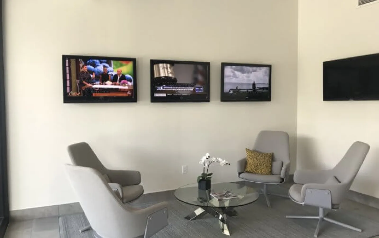 Modern waiting area with four gray chairs around a glass coffee table and three wall-mounted TVs.