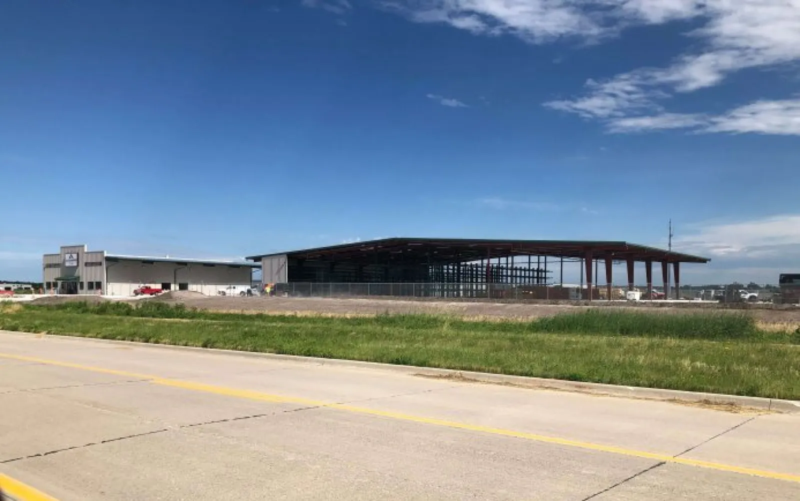 Construction site with steel frame building and adjacent warehouse under blue sky and scattered clouds