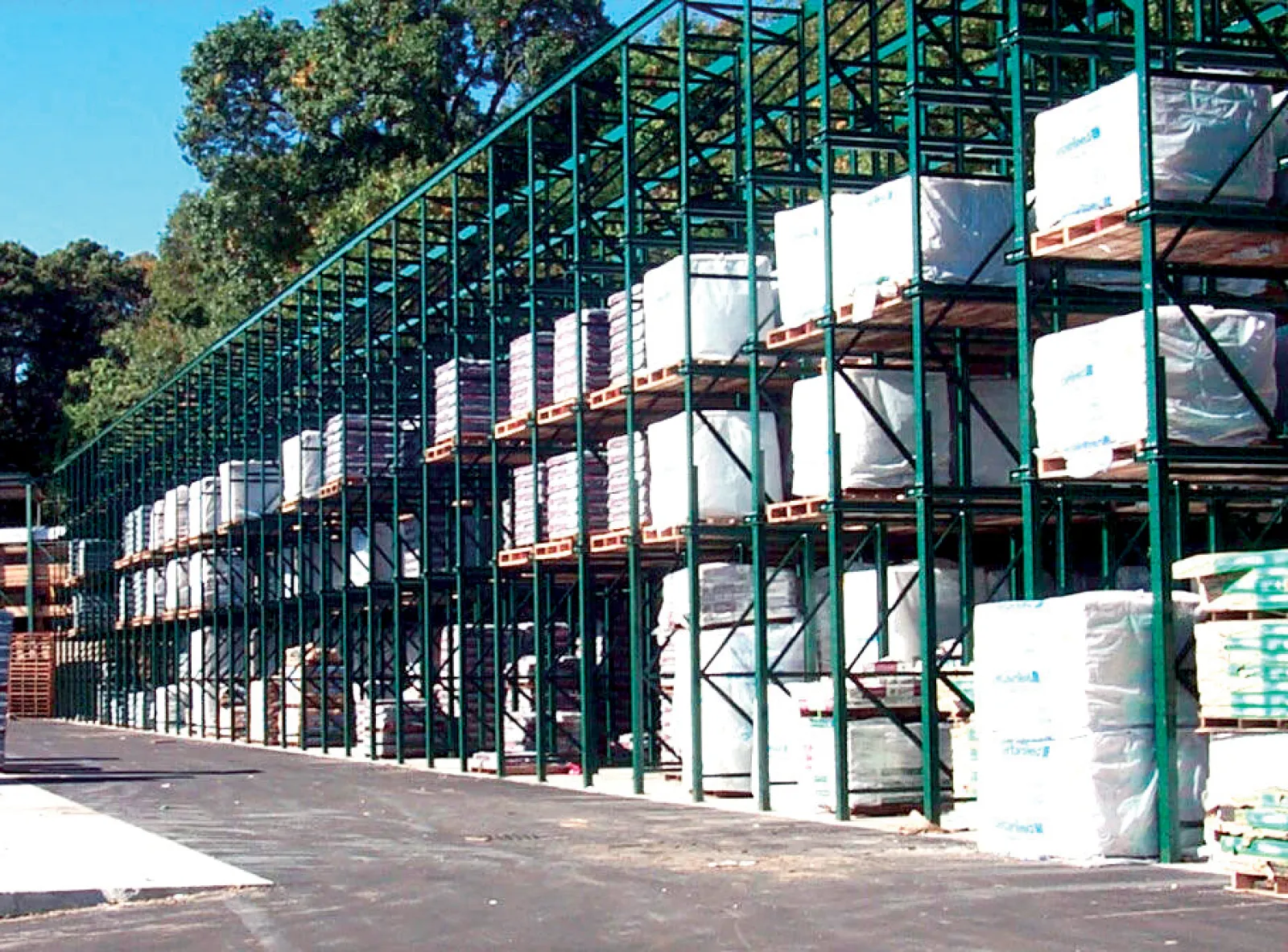 Outdoor warehouse racks filled with stacked pallets wrapped in protective covers under clear sky.