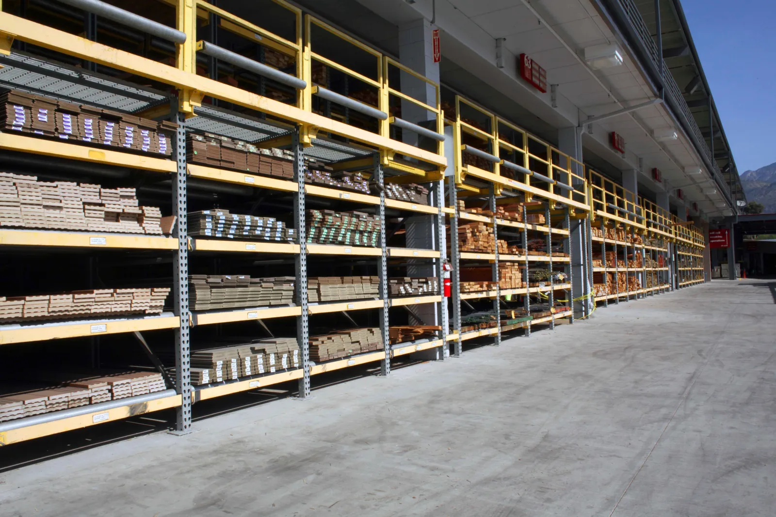 Outdoor lumber yard with organized wooden planks and beams stacked on metal shelving under a covered area.