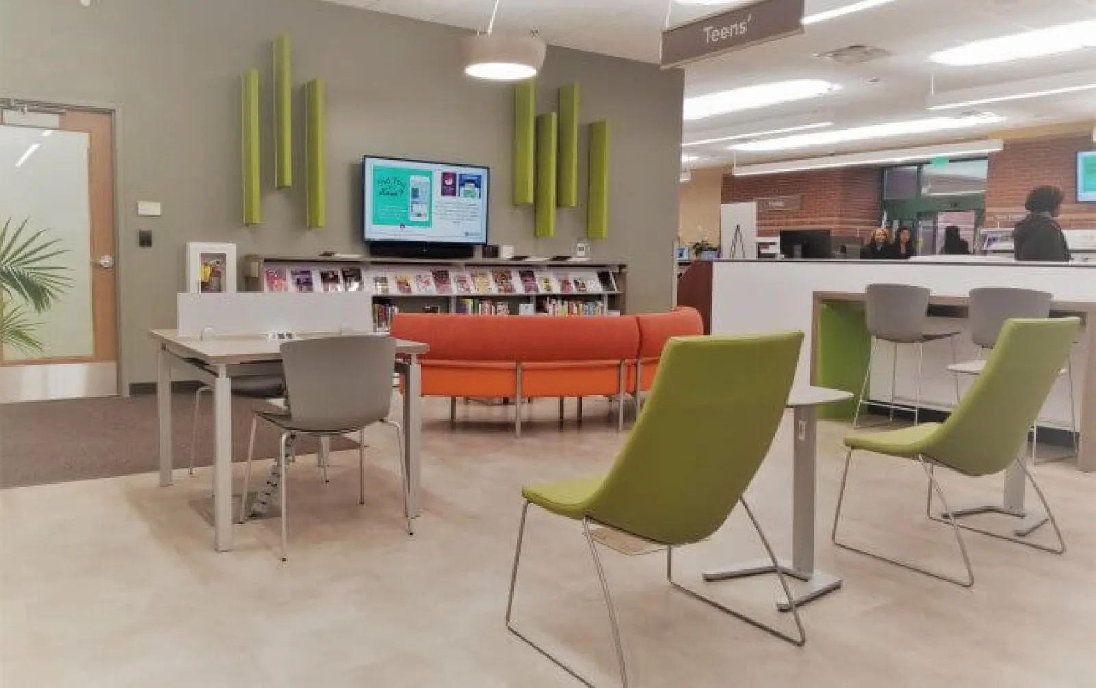 Modern library teen area with green chairs, orange couch, tables, bookshelves, and digital information screen.