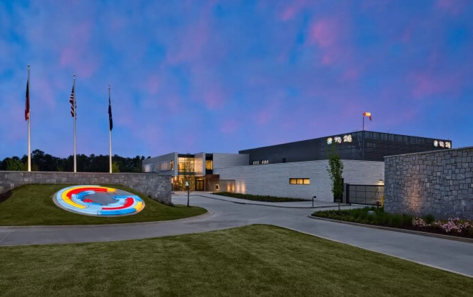 Modern building with illuminated circular sculpture and flags under a vibrant blue and pink evening sky.