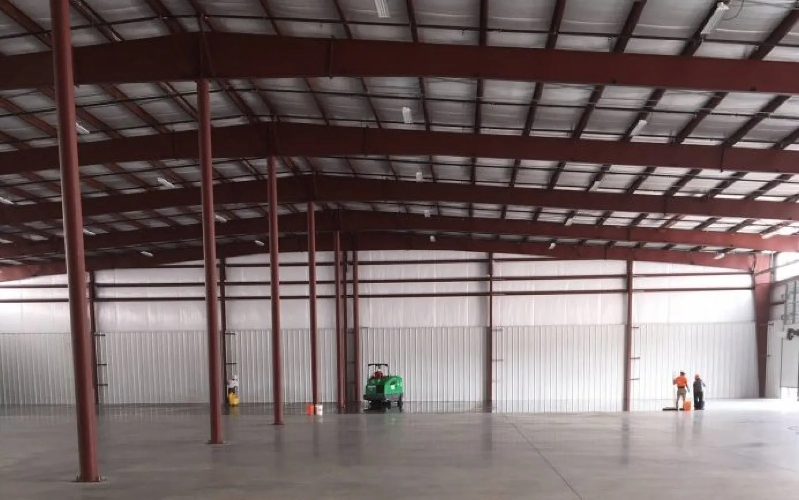 Large empty warehouse interior with steel beams, concrete floor, and two workers operating cleaning equipment.